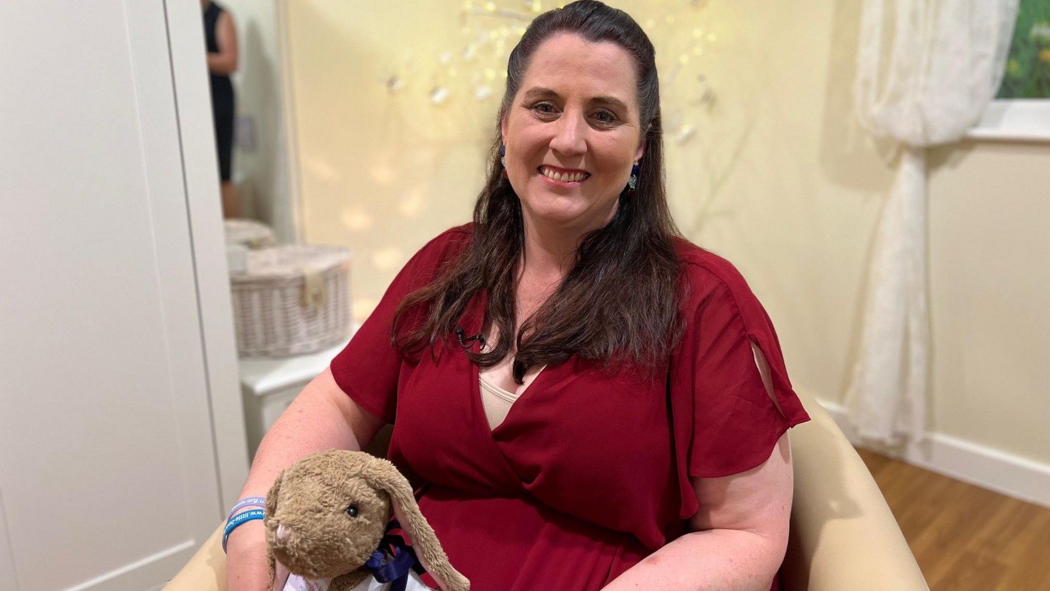 Ciara Curran wears a red blouse with her long brown hair partially tied back, holding a cuddly rabbit toy given out by her Little Heartbeats charity, with fairy lights in the background