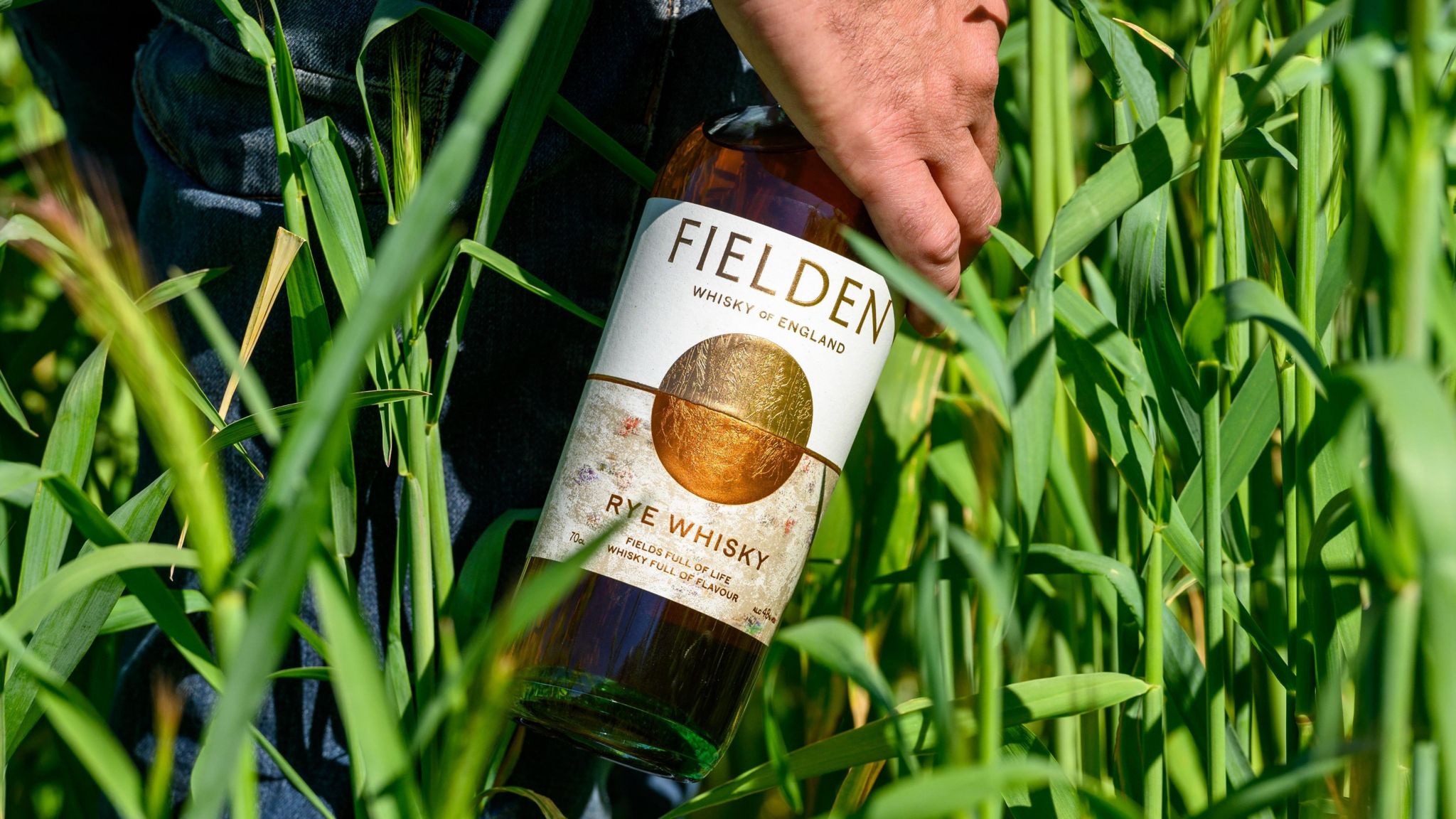 A hand holding a Fielden bottle in a field of wheat