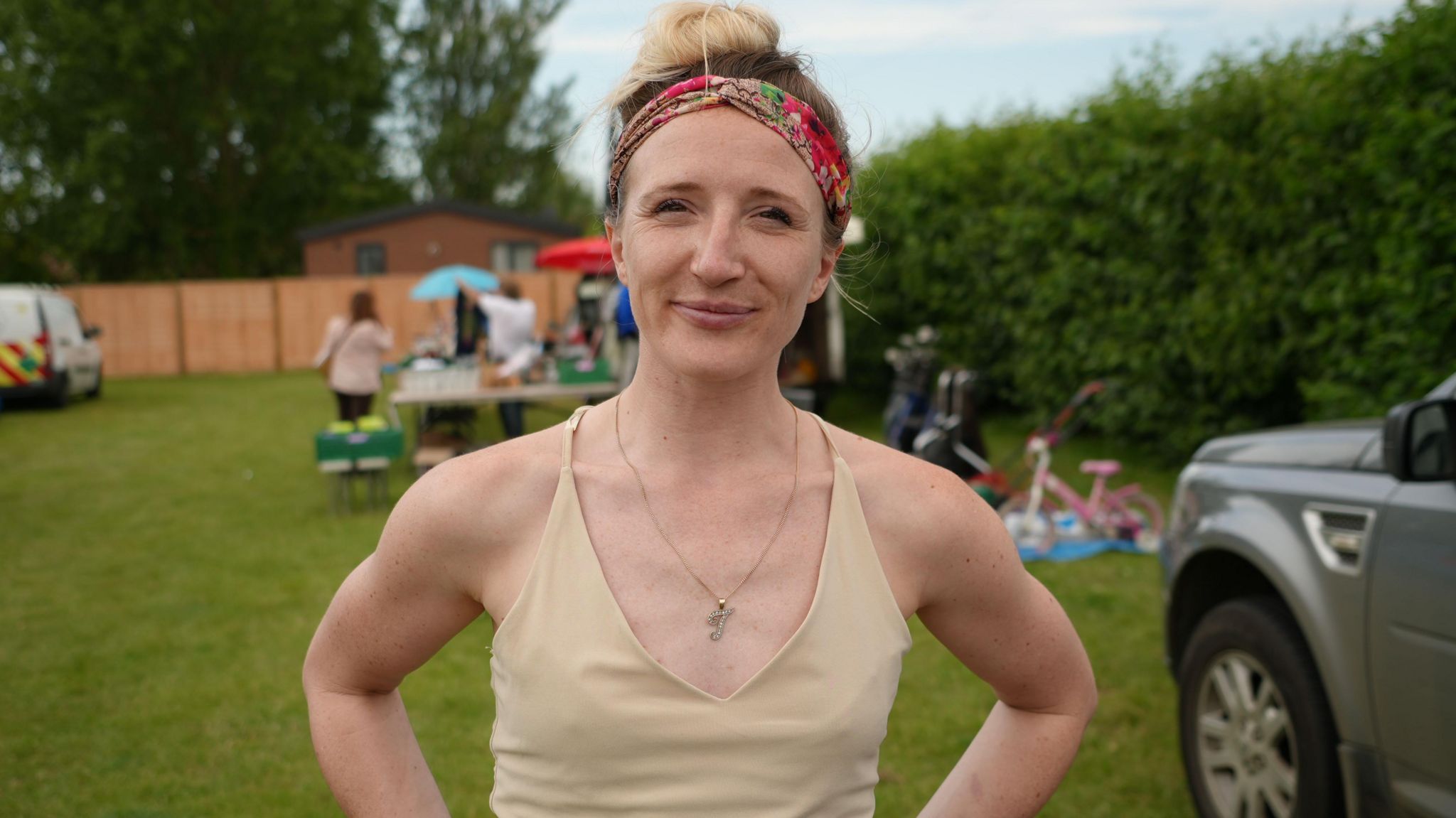 A woman with a cream top and colourful hairband - standing outside at a car boot sale