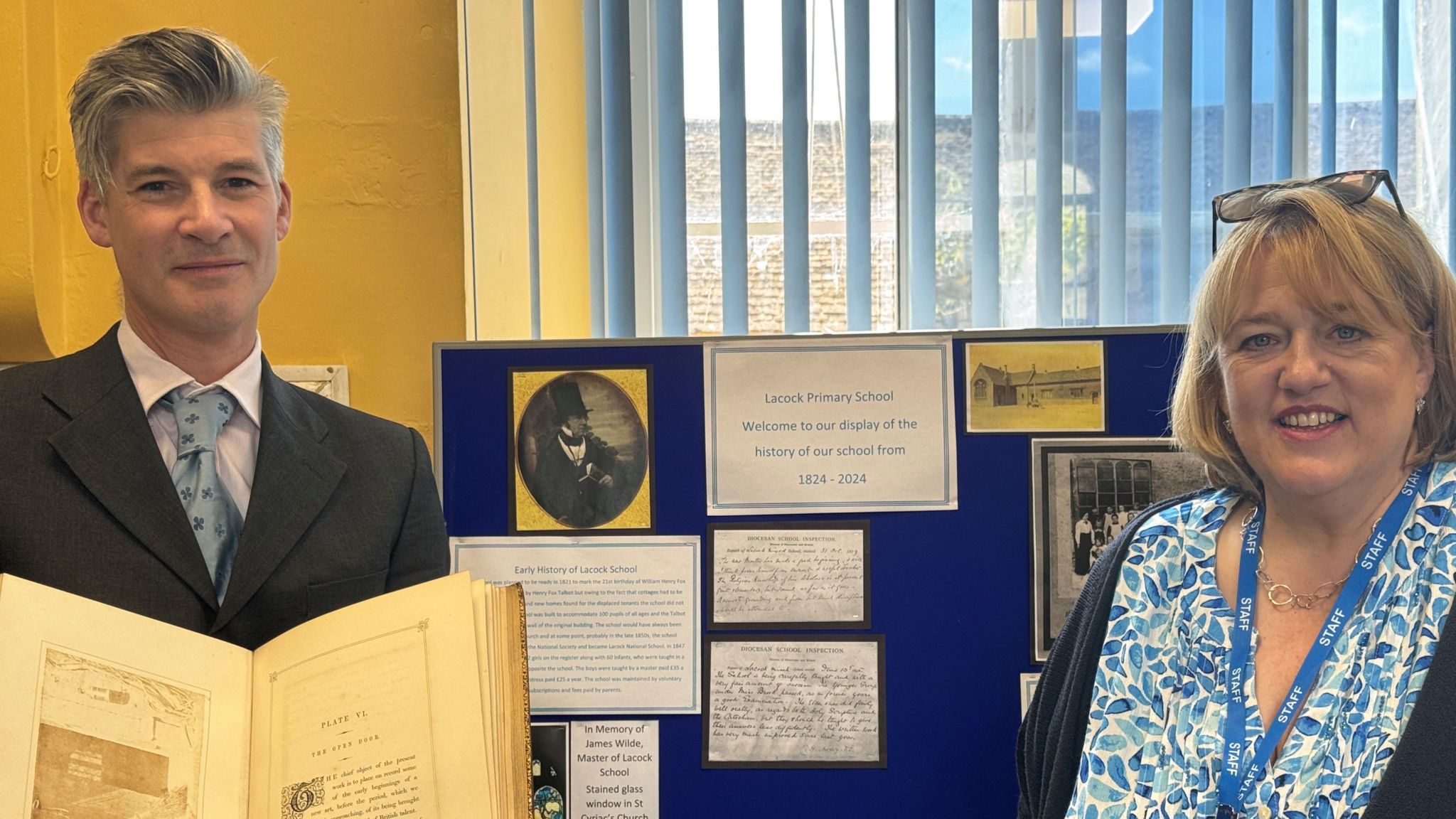 Revered James Clark-Maxwell holding a book while standing next to headteacher Caroline Jackson in front of an exhibition board at Lacock Primary School