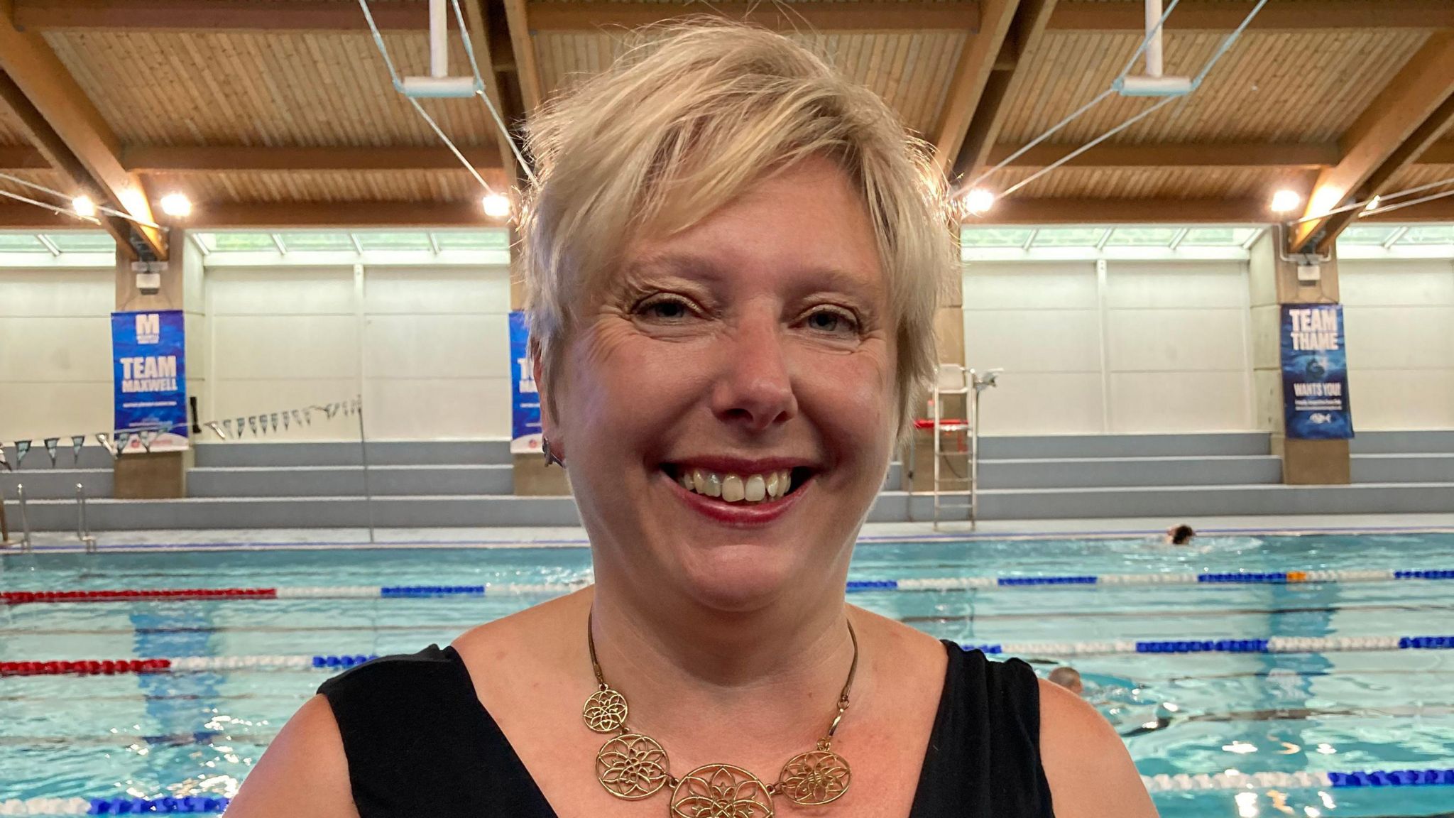 Emma Newman-Baronius, Olivia's mum, who has blonde hair and is wearing a black top and a necklace, smiles as she stands beside a swimming pool