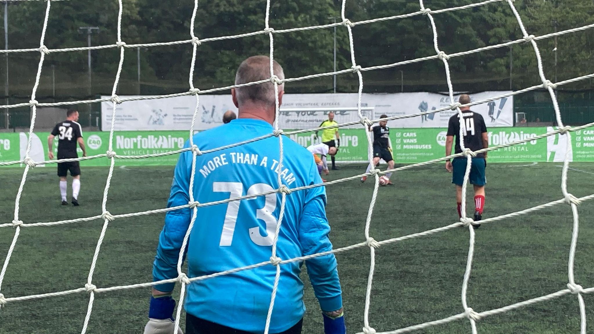 A football match from the viewpoint of behind the net of the goal. The goalie, who is wearing a light blue shirt that reads "More than a team" on its back, stands poised to receive the ball from players in front of him. 