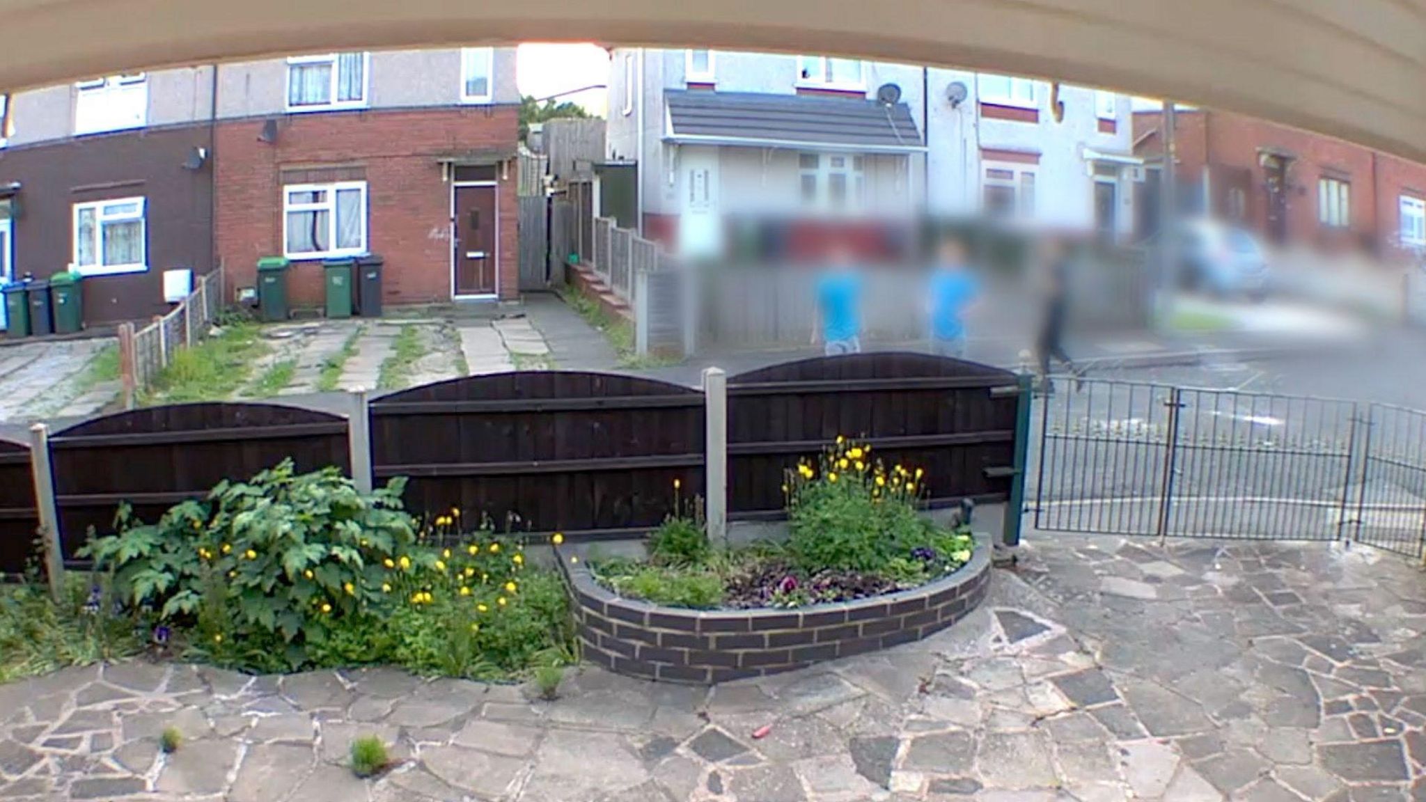 A picture of a front of garden opposite a row of houses, with three young people seen outside the property, who are blurred out