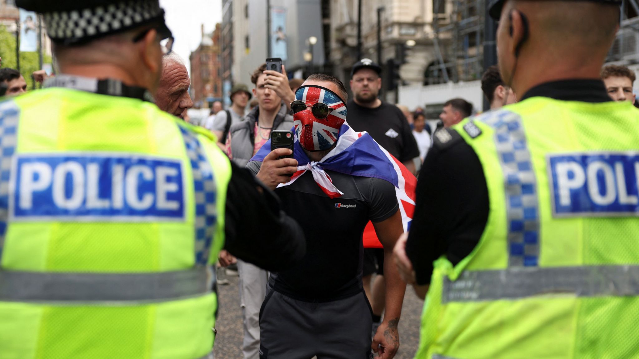 Two police officers watch as a man in a Union Jack mask films them with a mobile phone