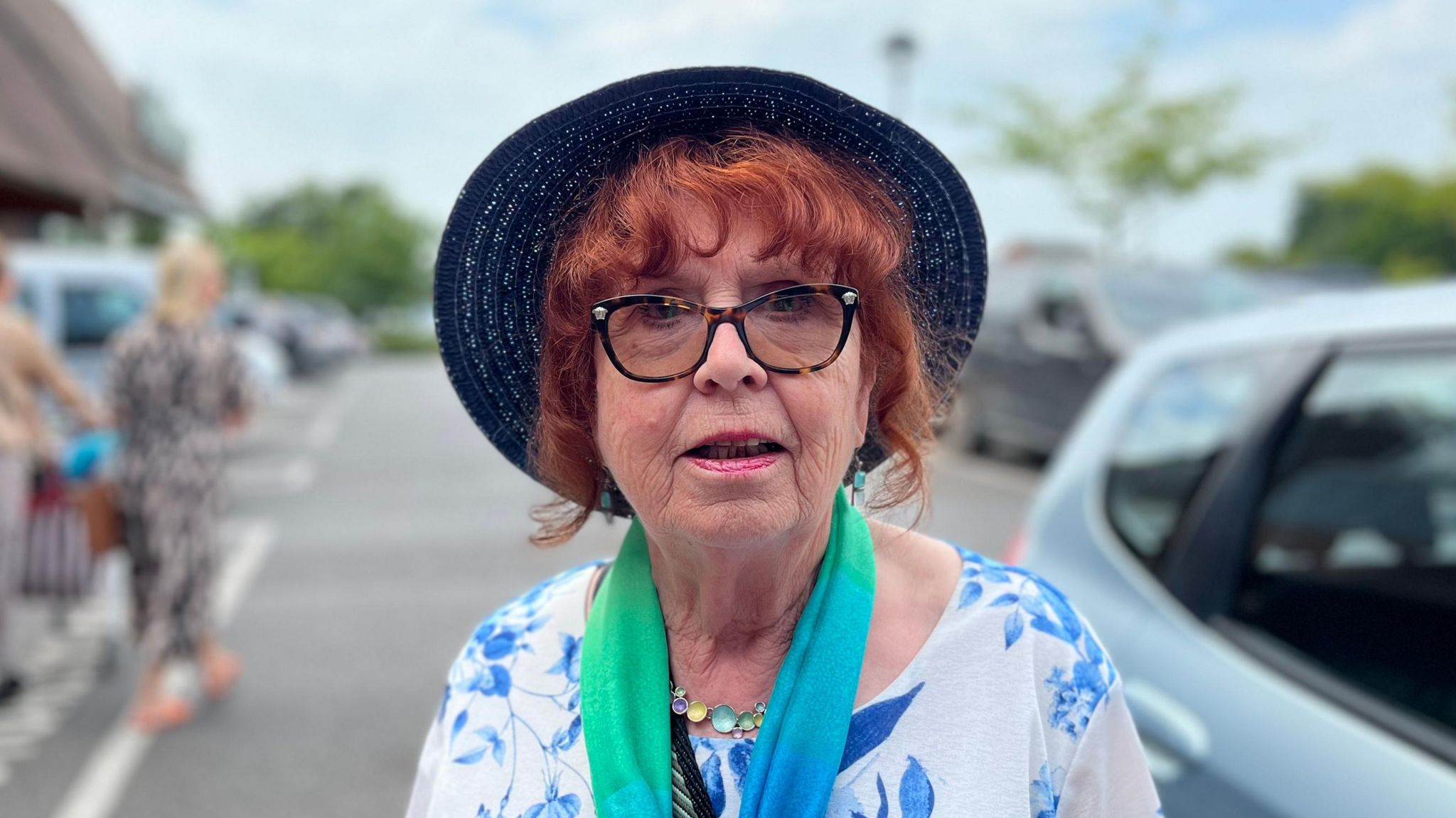 A woman looking at the camera. She has curly red hair and is wearing glasses with black frame. She is wearing a white top with blue flowers on it, with a green and blue scarf around her neck. The background is blurry but she is standing in a car park