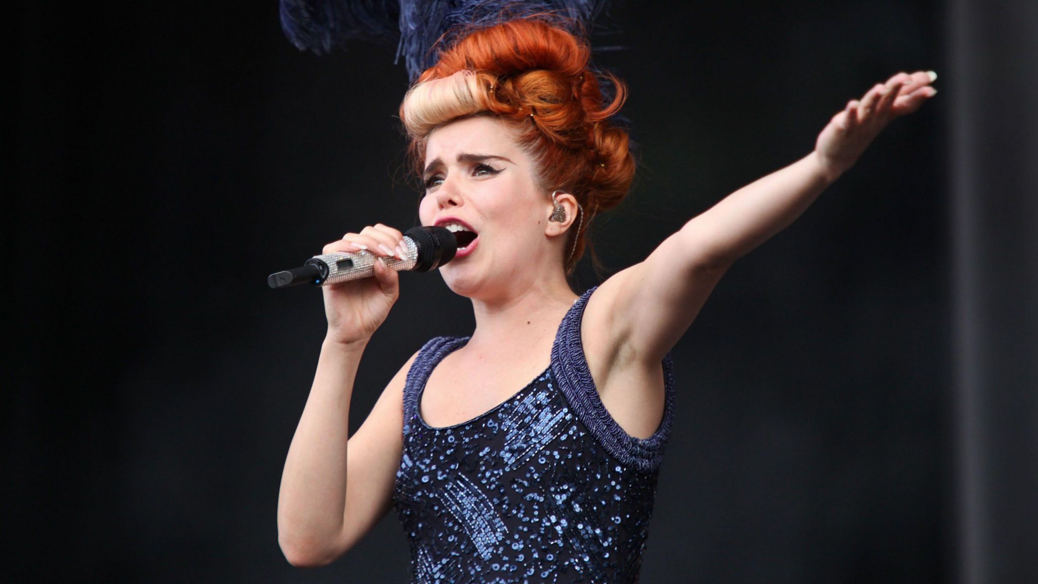 Paloma Faith singing on stage. She wears a sleeveless glittery blue top and has curled hair in shades of red and blonde