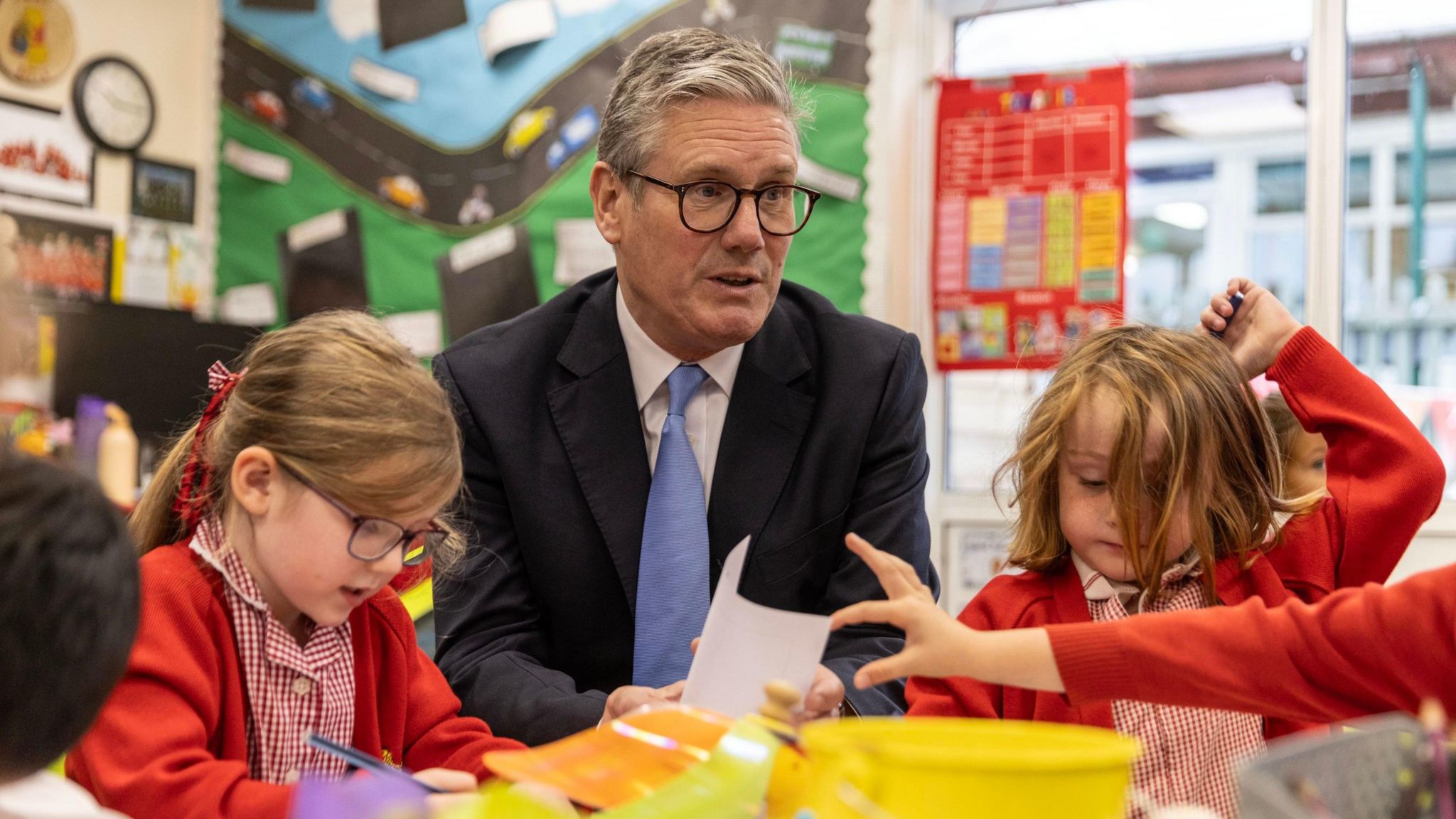 Prime Minister Keir Starmer, wearing a dark suit jacket and blue tie, sits at a primary school table between two girls in their school uniform, red cardigans over white and red shirts