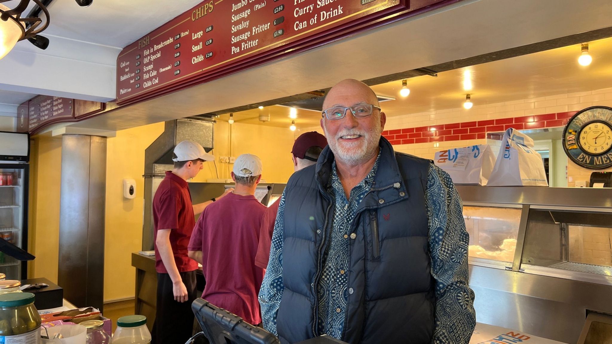 Fish and chip shop owner Jon Long standing behind the till wearing a navy blue body warmer and smiling at the camera while his staff work behind him