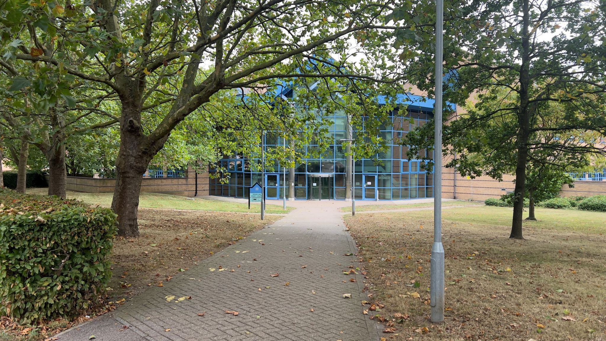 A path leading to Basildon Crown Court, which has a glass facade with blue edges to each panel. In the foreground is a brick path with trees and grass beside it.