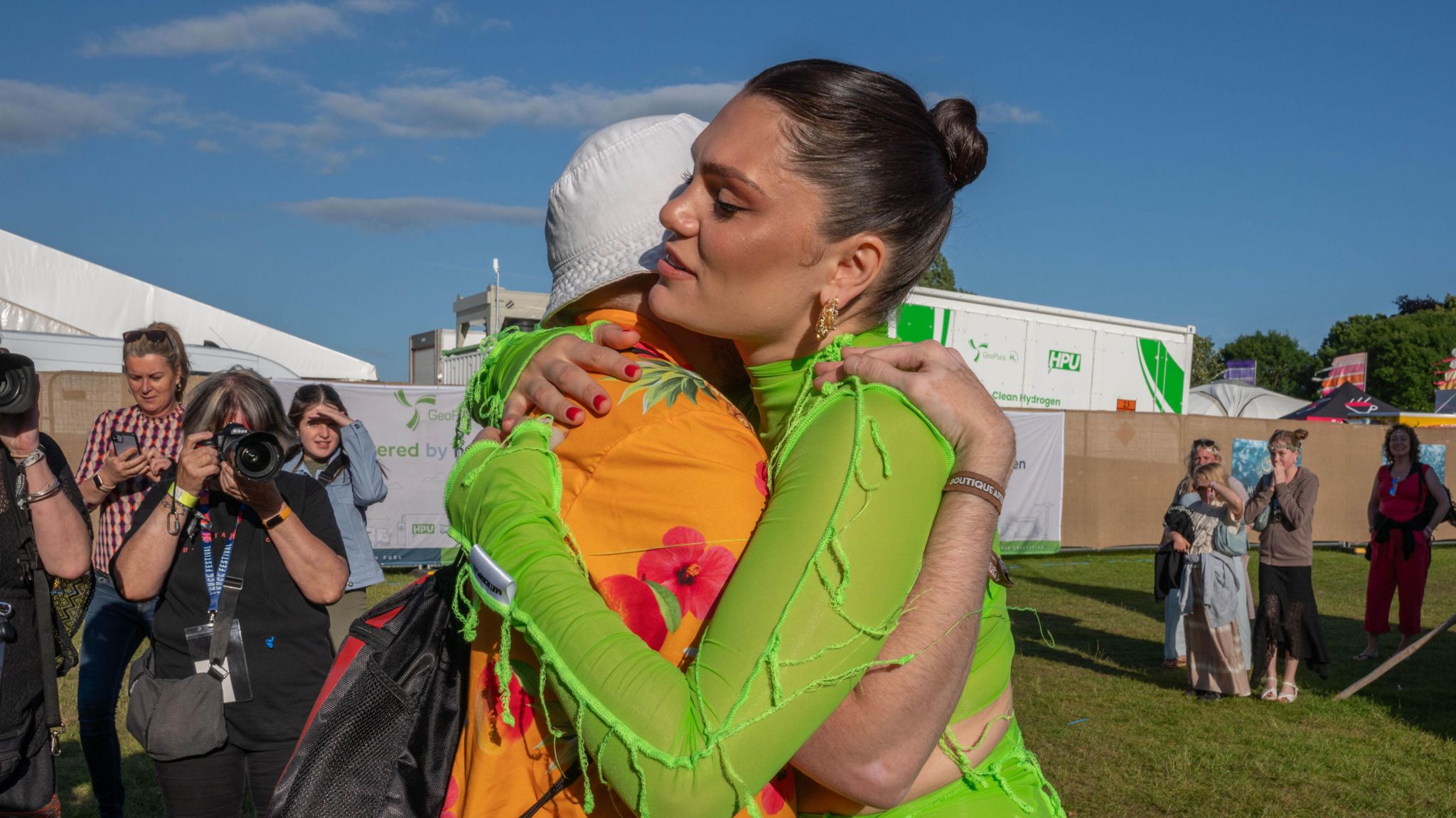 Jessie J in a lime green outfit hugging Nathan who is wearing a orange shirt with pink flowers on. He is also wearing a white bucket hat and crowds of photographers and spectators are around watching the embrace