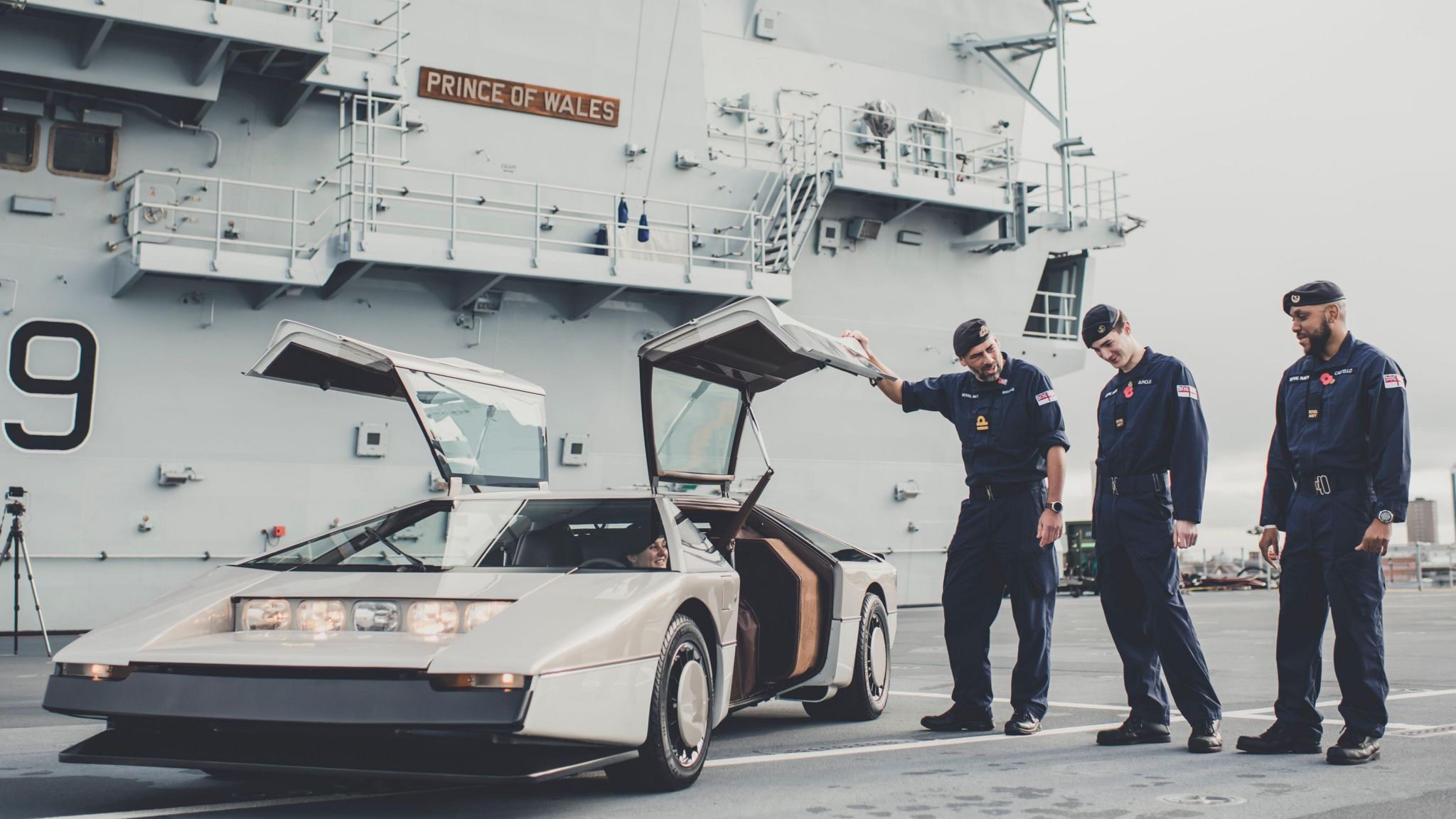 A cream and beige supercar with an angular shape and pointed nose is parked in front of a huge military ship. Three Navy personnel in blue uniforms stand in front of it.