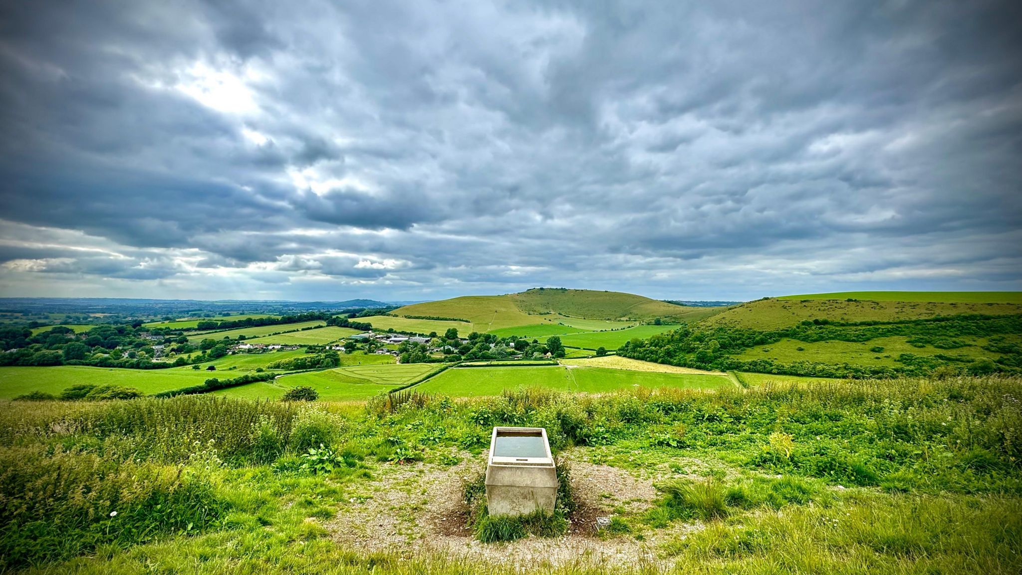 THURSDAY - The rolling green Dorset countryside is photographed from Compton Abbas. A metal water trough is in the centre of the frame, behind the countryside is dotted with trees and hedges with two hills. The sky is covered in grey clouds