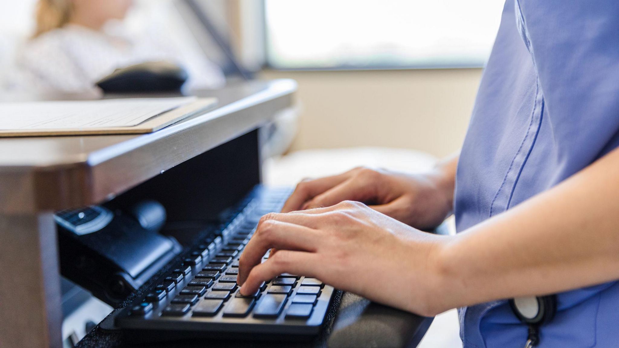 A healthcare worker in blue scrubs types on a keyboard