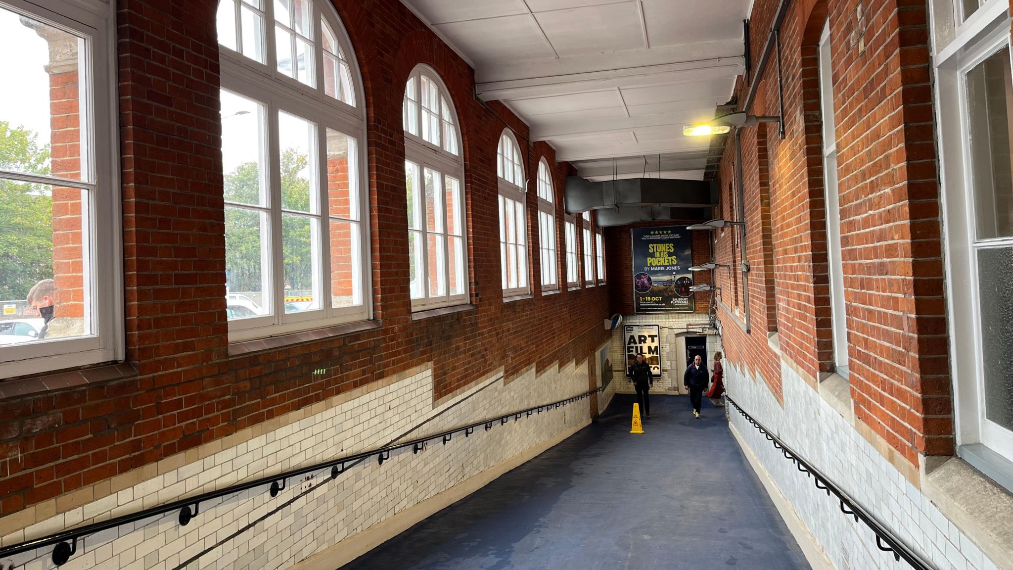 The slope into a subway, historic metro-style tiles in white down the side with black railings and large, arched windows before it sinks underground