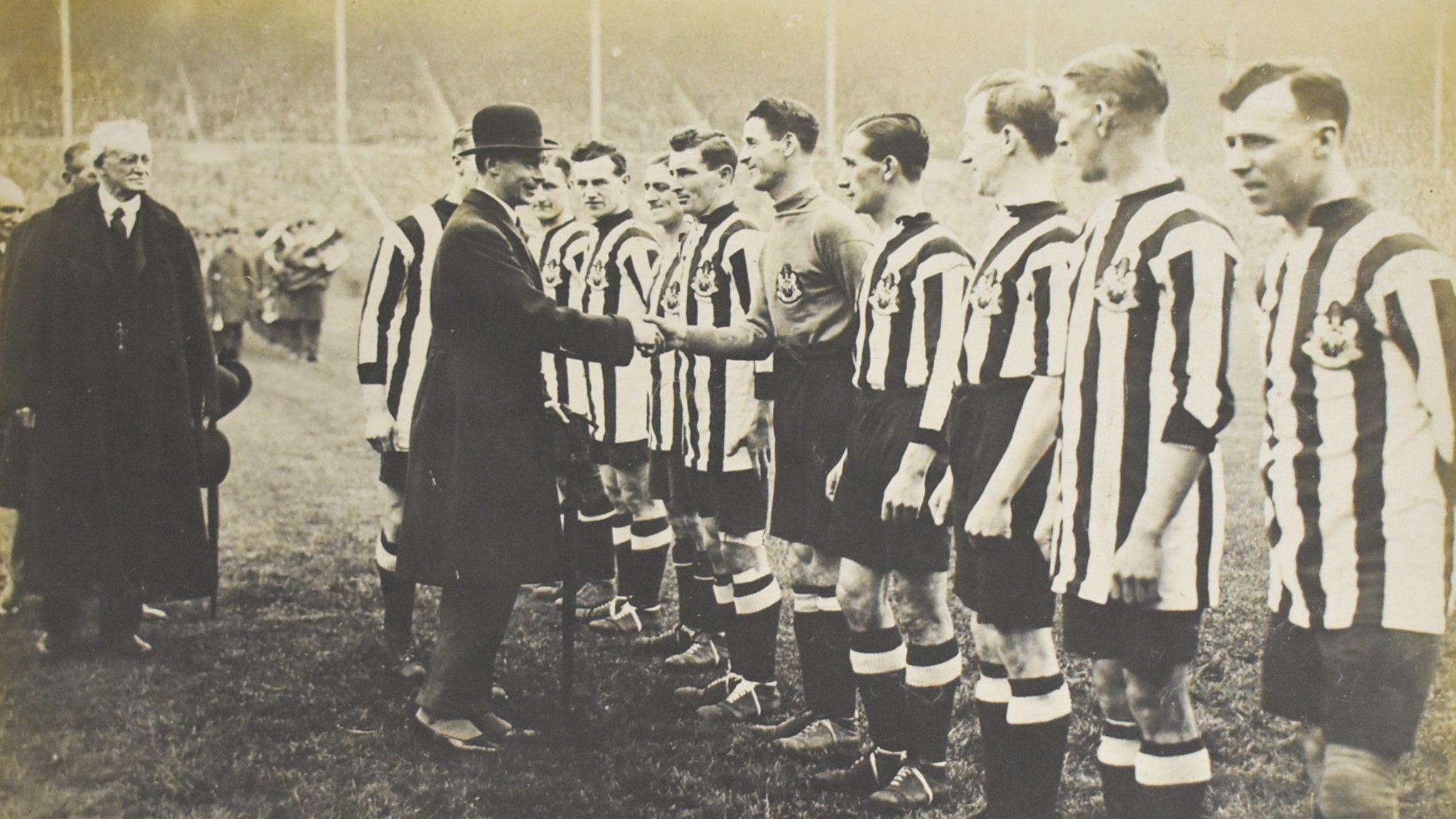 Players stand in a line in Newcastle black and white kit. The king is in a bowler hat and shakes one of the player's hand.