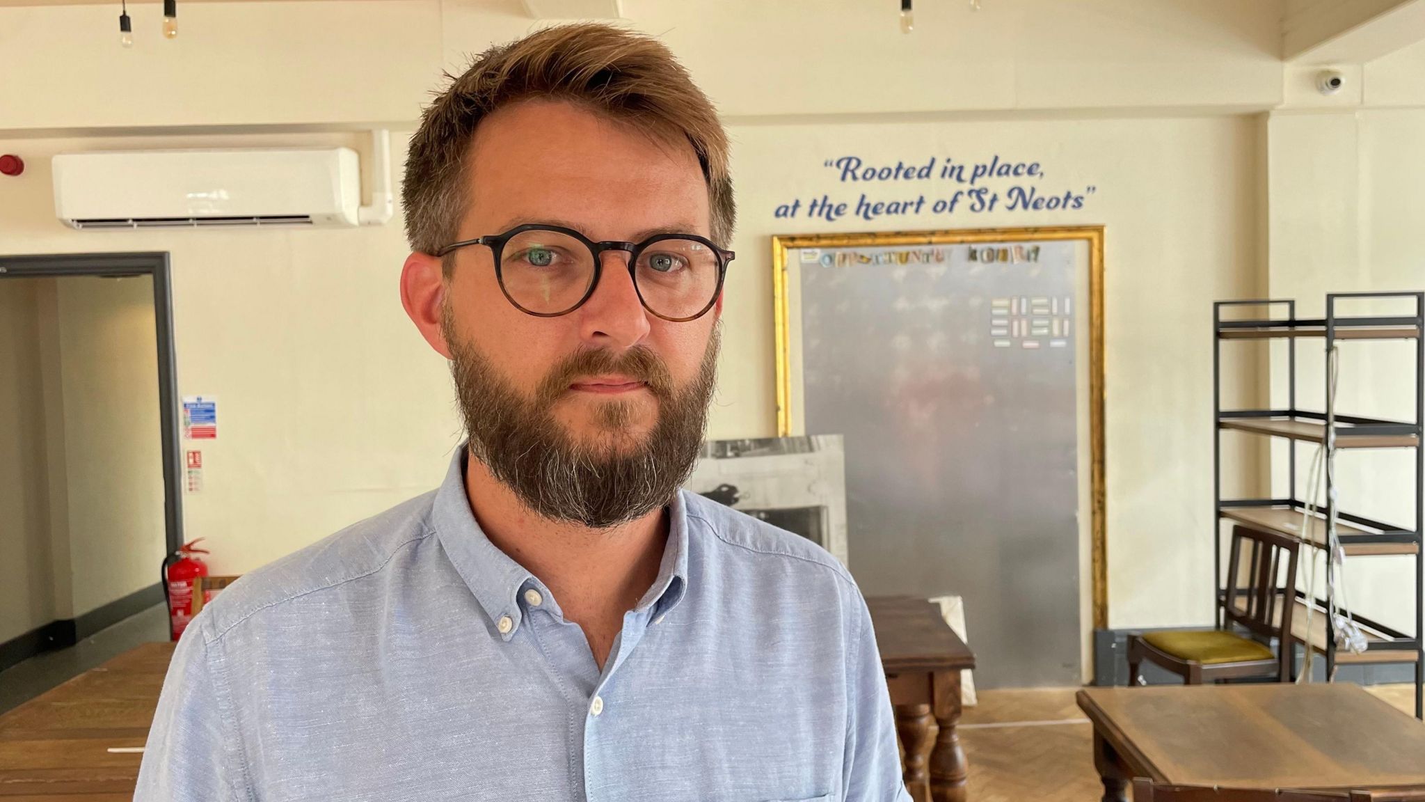 A man, Alex Hughes, is wearing glasses, a blue shirt and has a beard. At the back of the room he's standing in is a sign which says 'rooted in place, at the heart of St Neots'