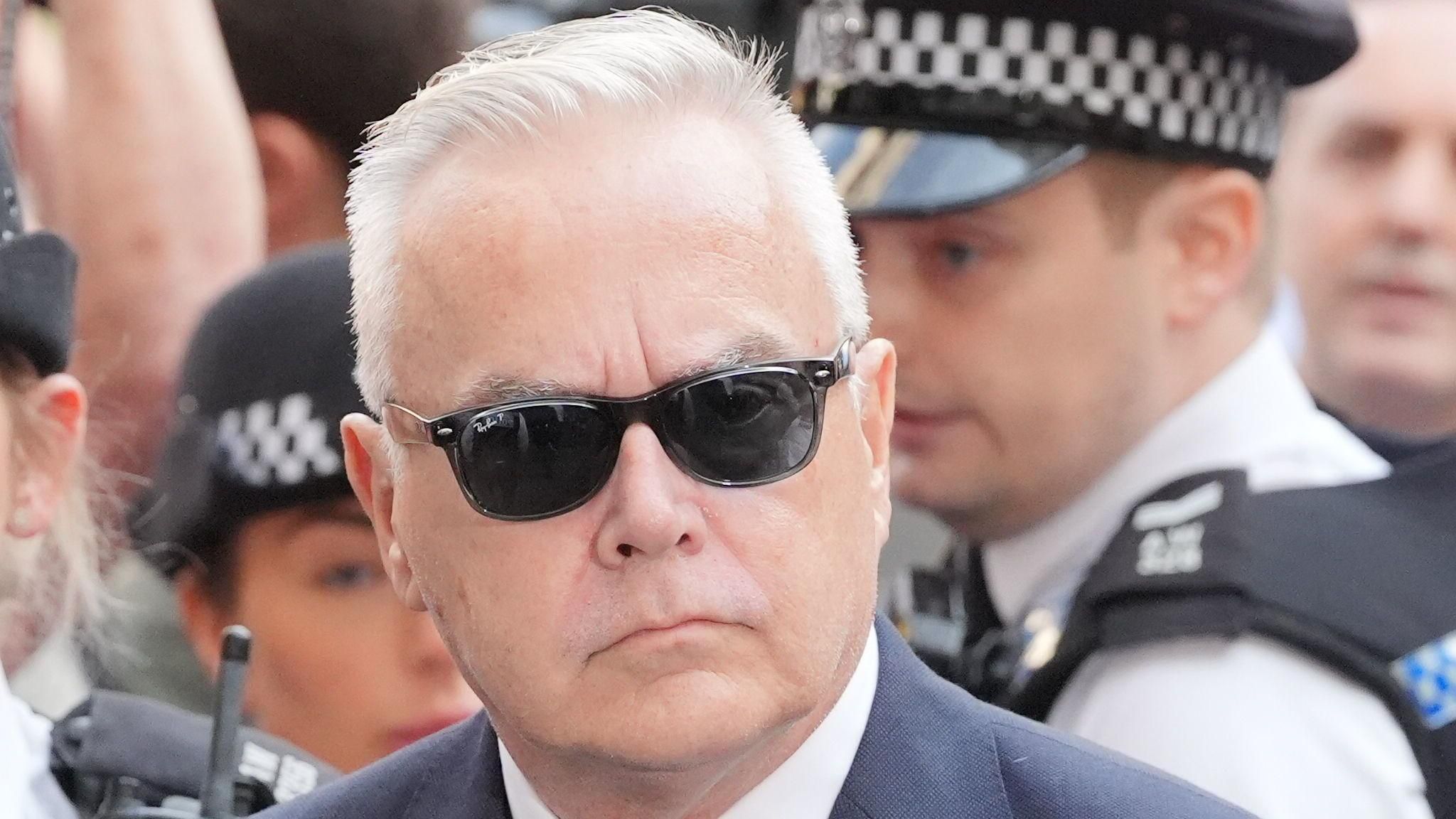 Huw Edwards outside court, wearing suit and dark sunglasses, accompanied by police 