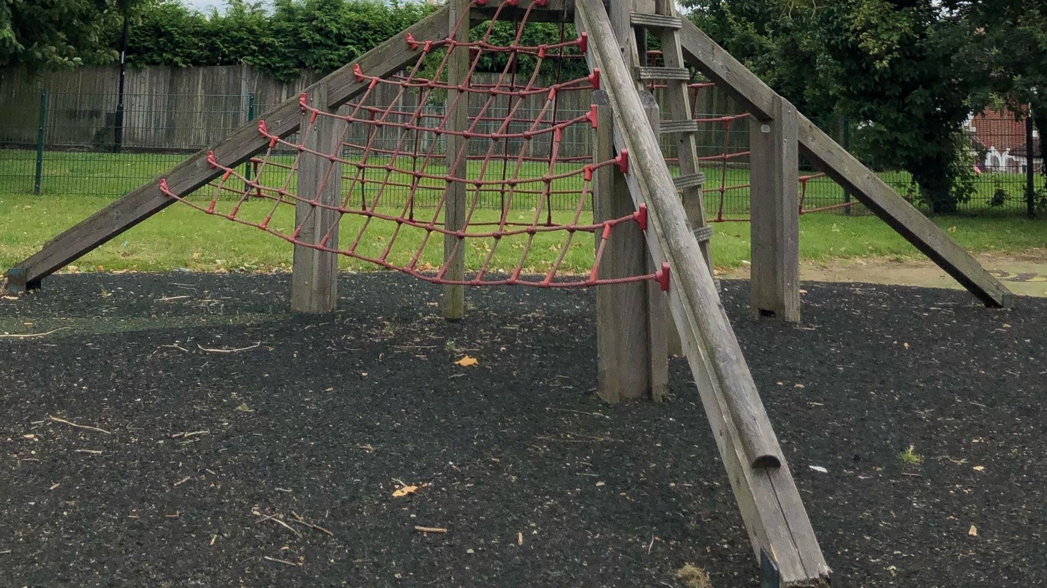 Two children perch on the top of a wooden climbing frame in a park. On one side of the climbing frame there is a large rope ladder. In the background there is grass and trees.