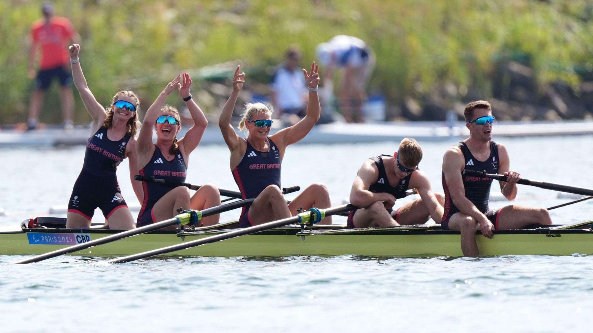 Five rowers celebrate in their boat after winning their final at the Paris 2024 Paralmpics. The crew of three women have their arms aloft, while the two men at the rear of the boat are recovering from their efforts.