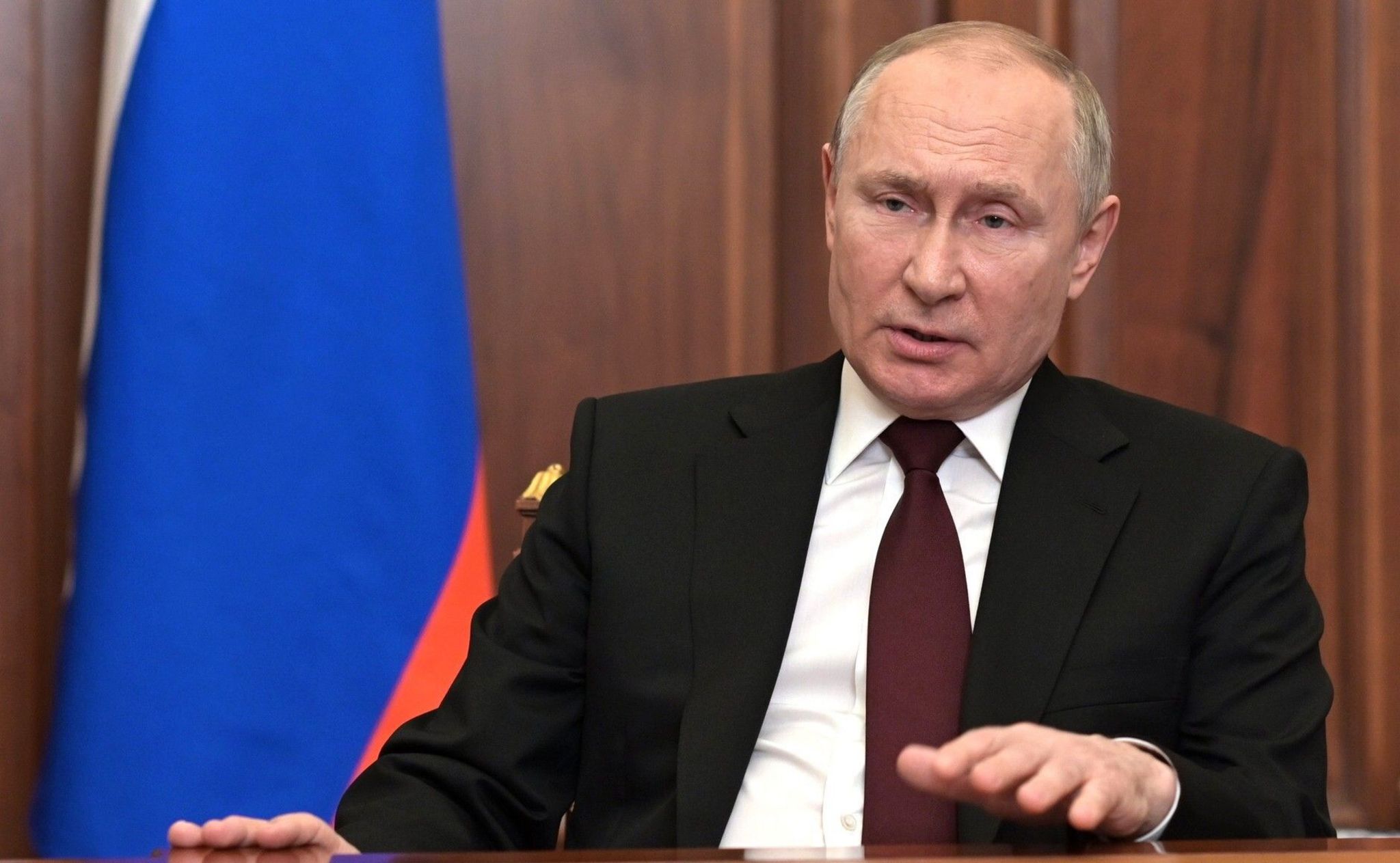 Vladimir Putin pictured delivering an TV address in Feb 2022 as he announced Russia's invasion into Ukraine. He is wearing a suit and is sitting in a wood-panelled room with a Russian flag to the left of the photo.