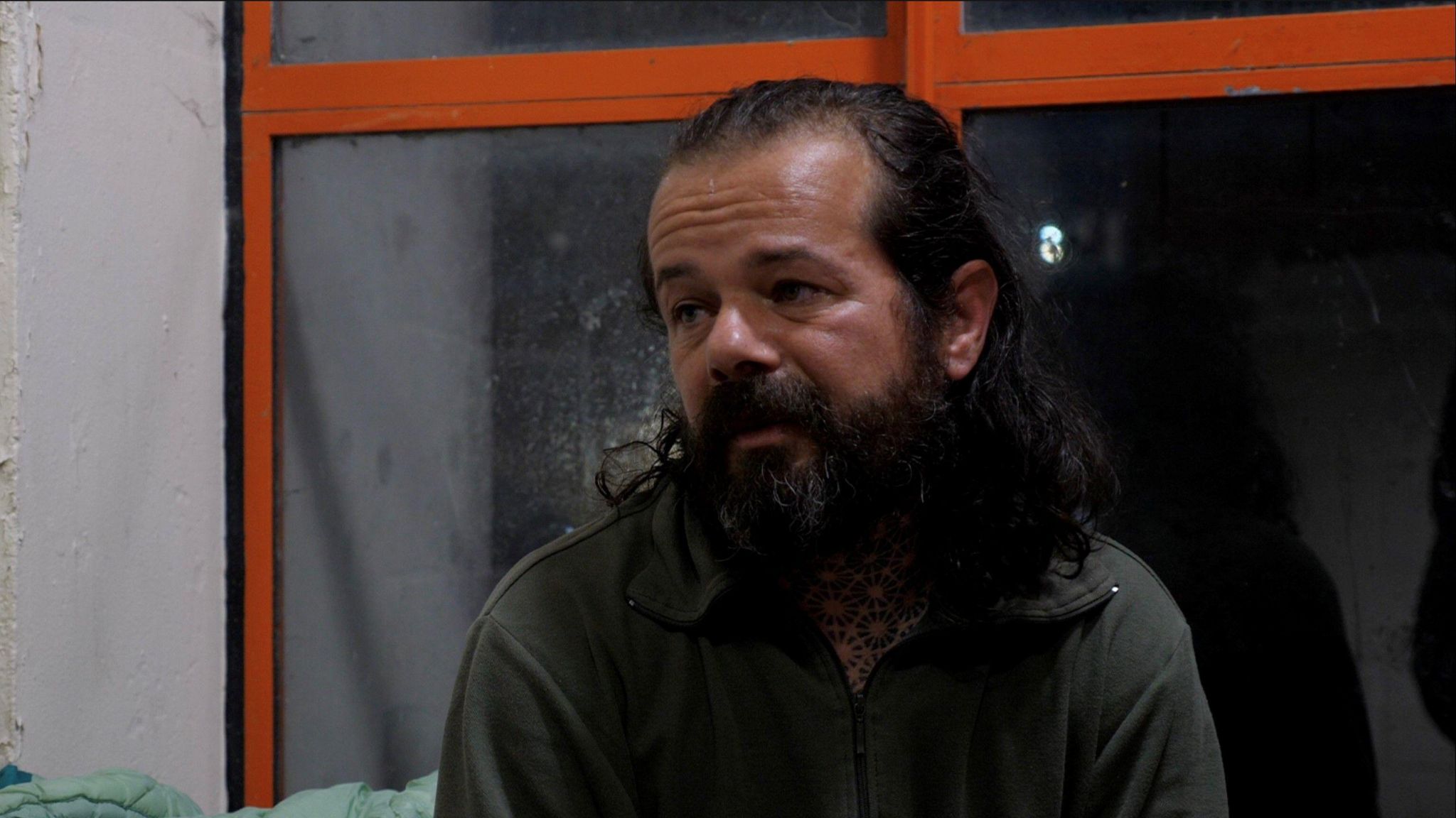 Jakab, who has long hair and a thick beard, sits in front of a tinted window with red frames. He is looking off camera to the left and wearing a dark green zip-up top