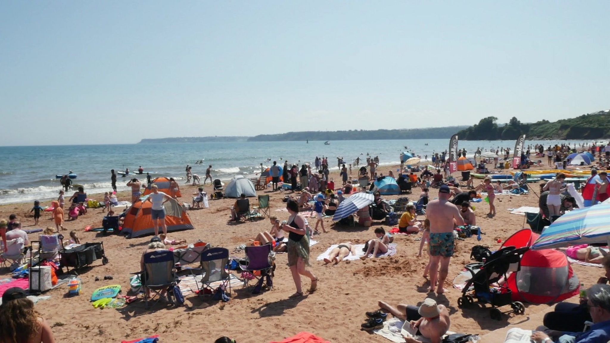 A photo which shows Goodrington Sands in Paignton. Hundreds of people are on the sand with deck chairs, towels and tents, as well as umbrellas. Some people are sunbathing