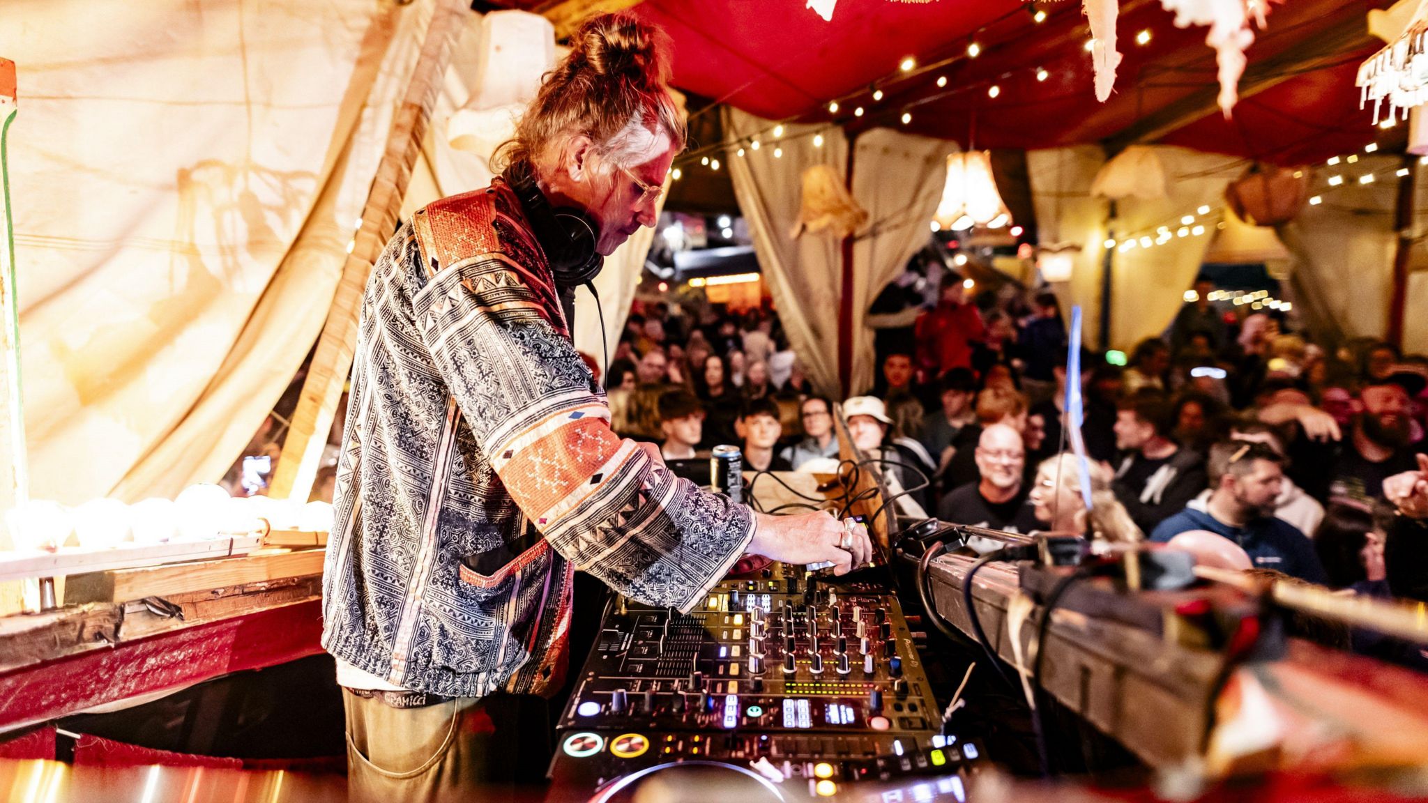 A man with glasses and his hair in a bun stands in front of a black DJ deck above a crowd. He is pushing buttons. He is inside a beige-coloured tent, and below him is a crowd of hundreds of people