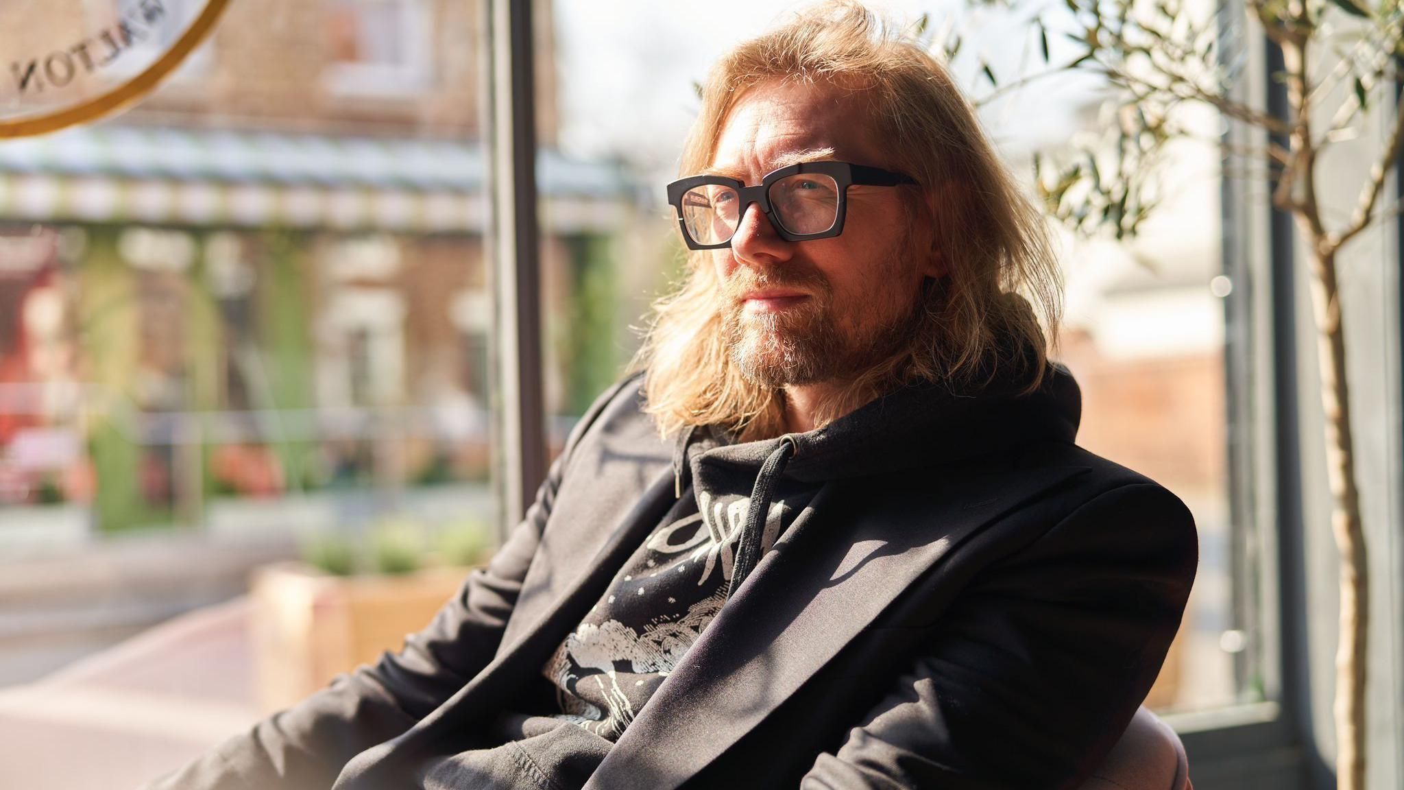 Paul Southouse, Founder of Visit Jericho, sat on a chair by the window of his LYNRACE MMXXI bar. His mid-length blonde hair is down and he is wearing black framed glasses and a black blazer.
