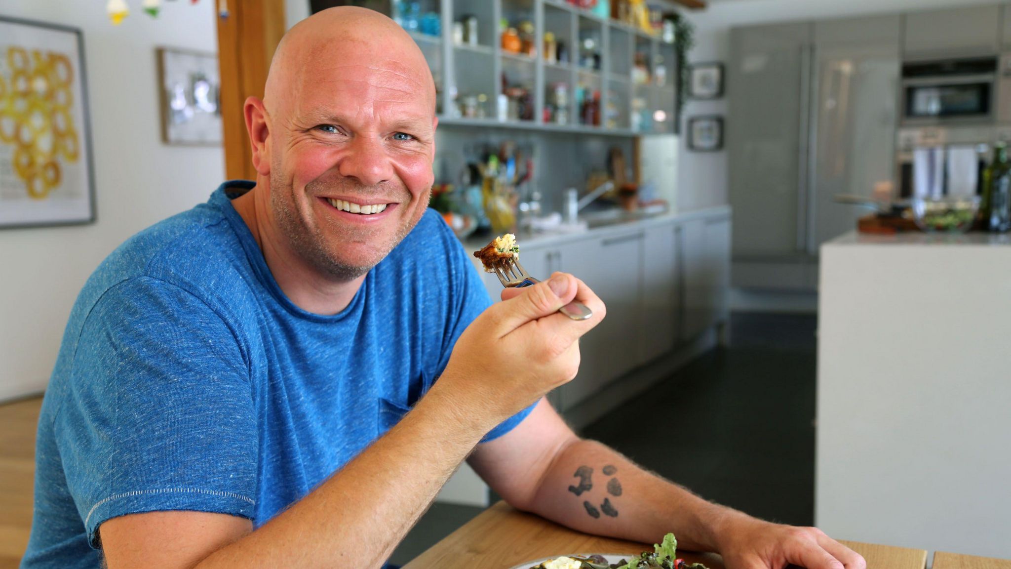 Tom Kerridge sat at a table, eating and smiling