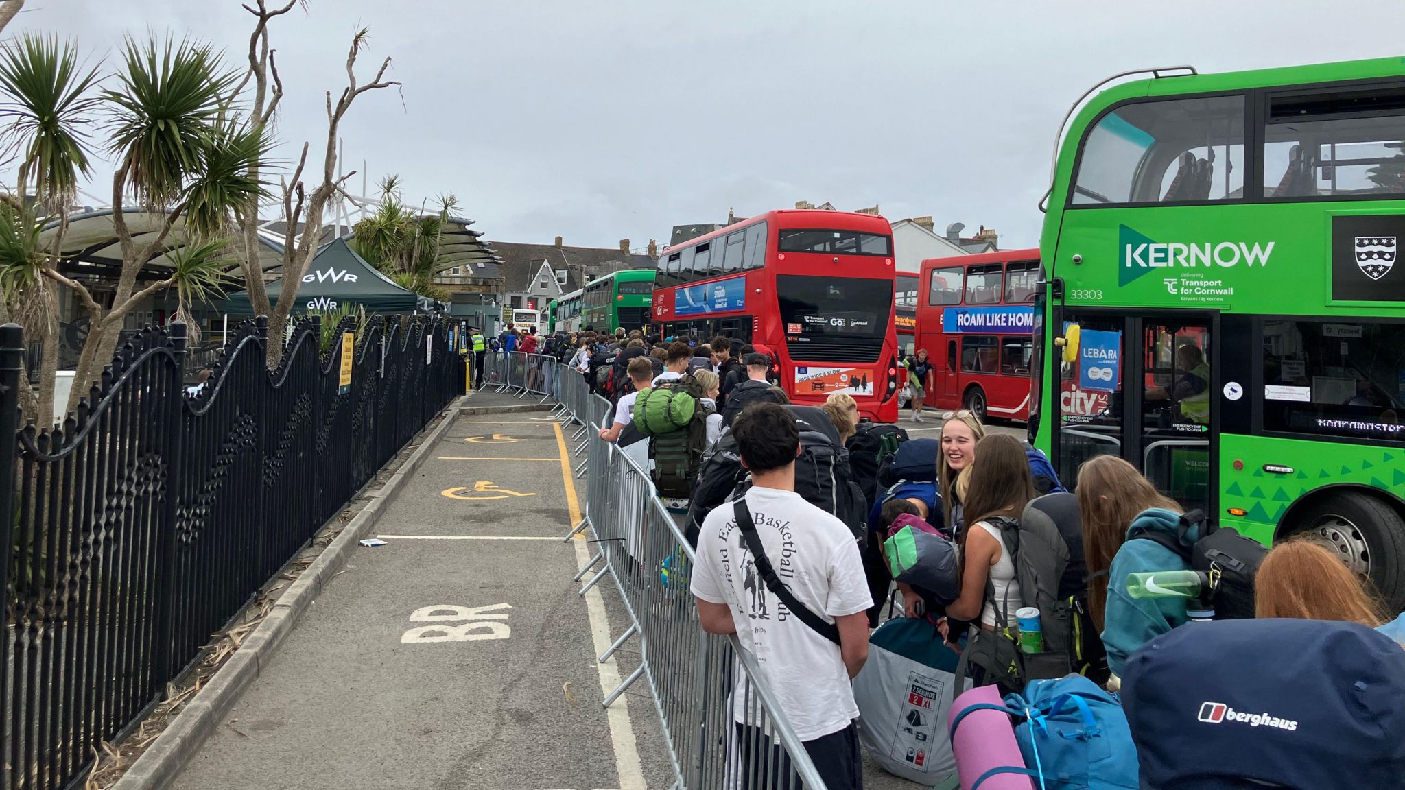 People queuing to get into Boardmasters festival. A line of people are on the right hand side of the image next to a metal barrier. In the background there are three Cornwall buses