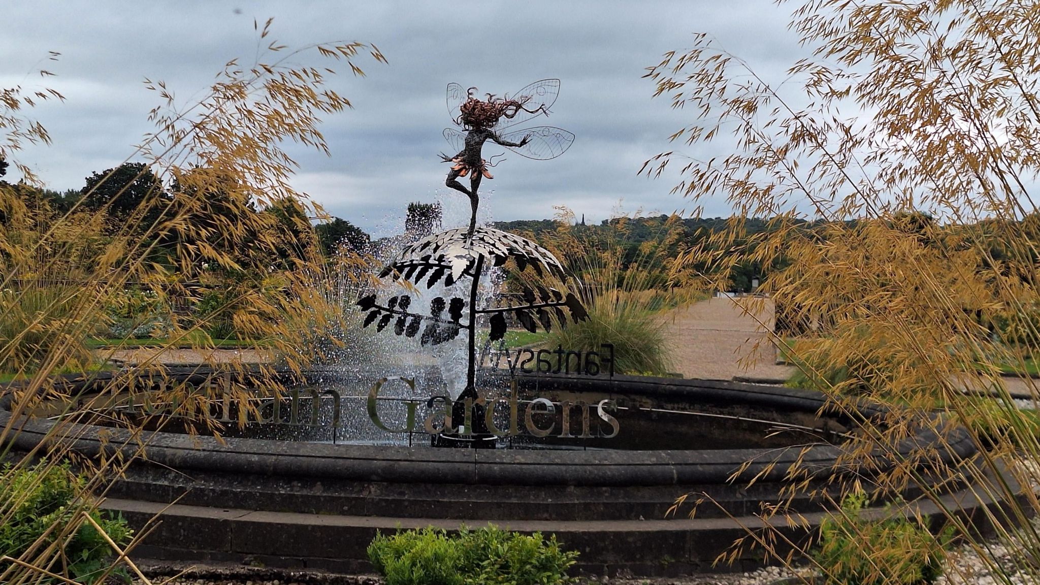 A metal sculpture of a fairy set against a water feature, with plants and grasses encroaching the foreground from the sides. 