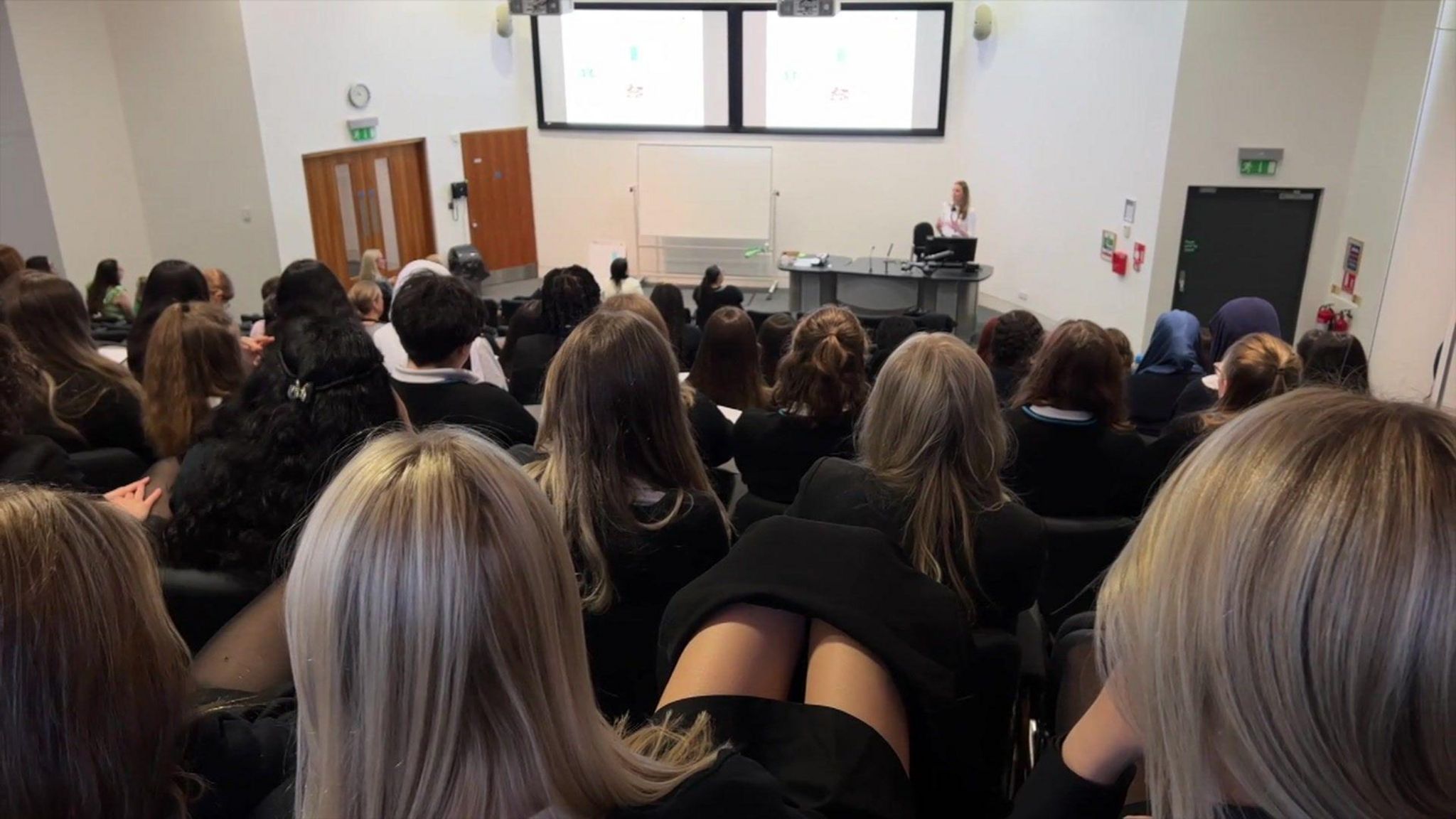 Around 30 young female school students sitting in a lecture theatre listening to a talk from a middle-aged woman