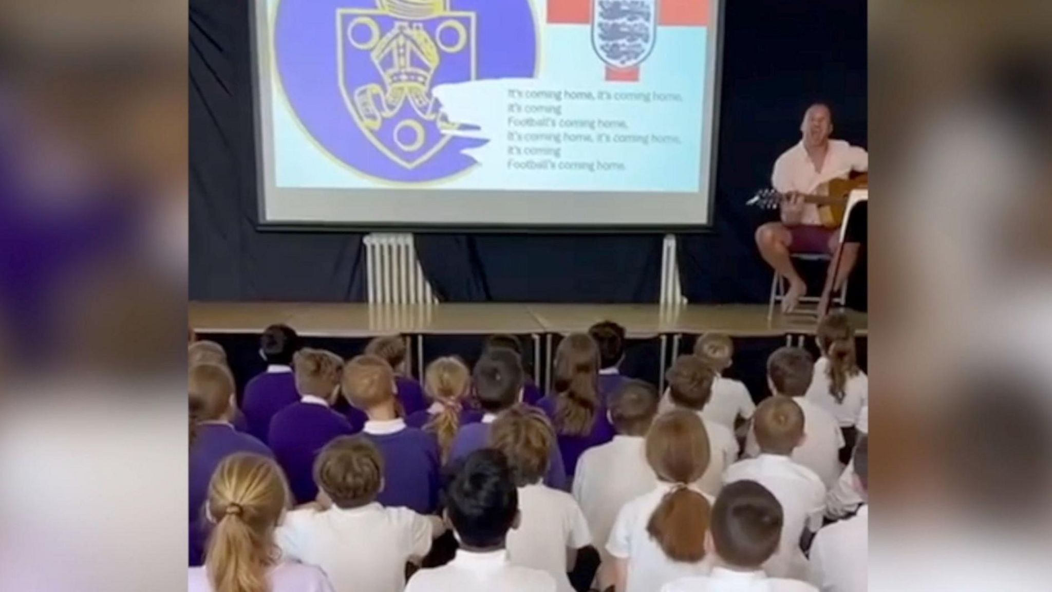 Still taken from the video released by the school of pupils sitting in an assembly hall facing towards a stage singing, the headteacher, Mr Spooner, is sat on the stage with a guitar next to a projector screen which has the words to the song on it, specifically the chorus 'It's coming home'