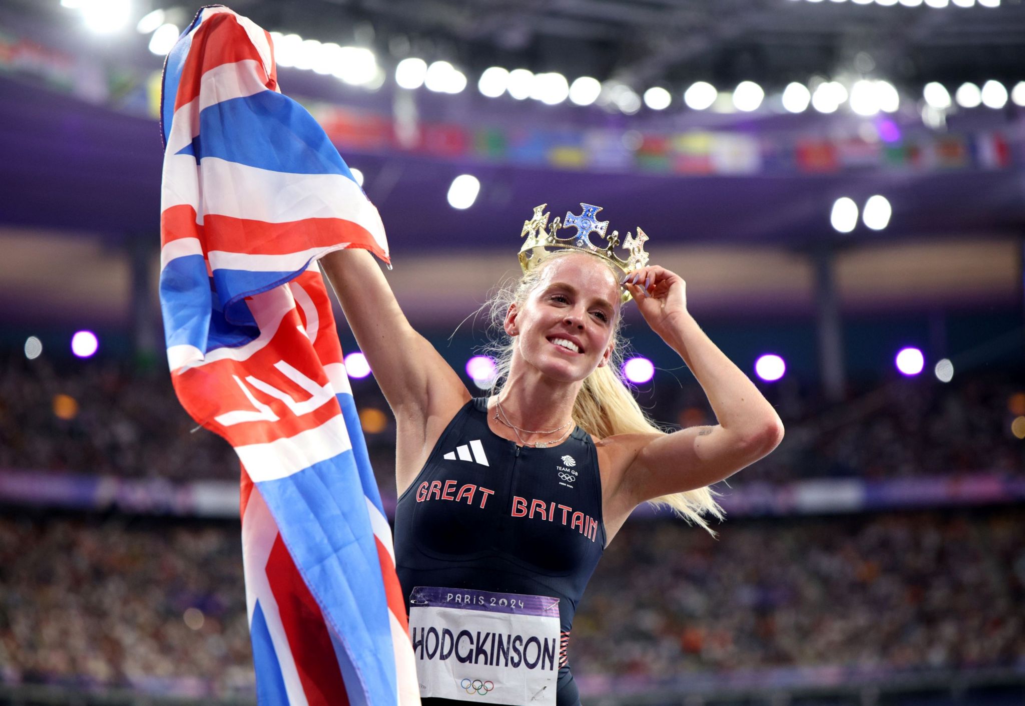 Keely Hodgkinson holds a Union Jack flag in one hand and a crown on her head in the other. She is wearing a dark blue vest with Great Britain and her name on. The crowd behind her is blurred.