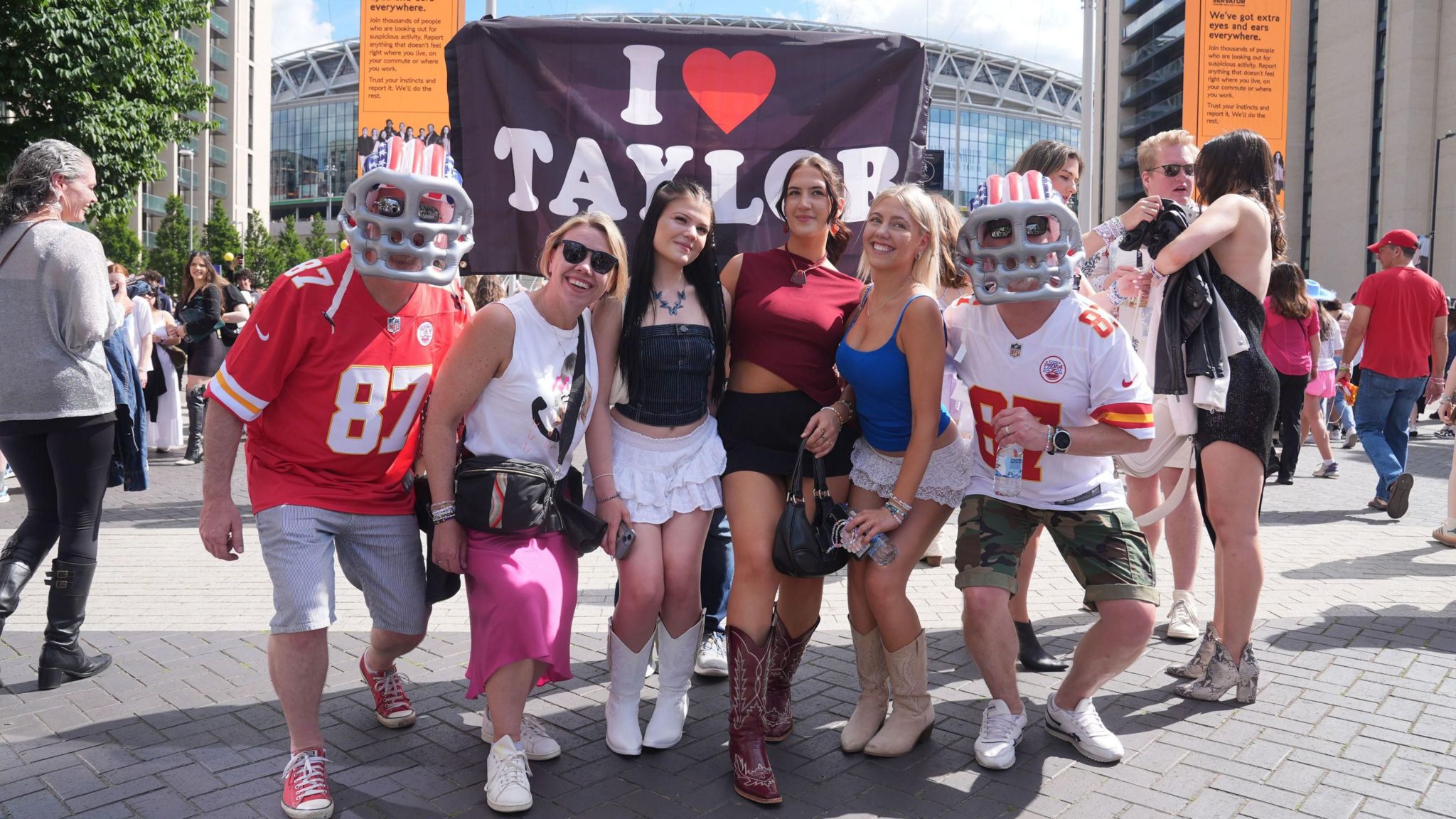 Fans in cowboy boots and American football costumes outside Wembley, with a banner that says I Love Taylor above them
