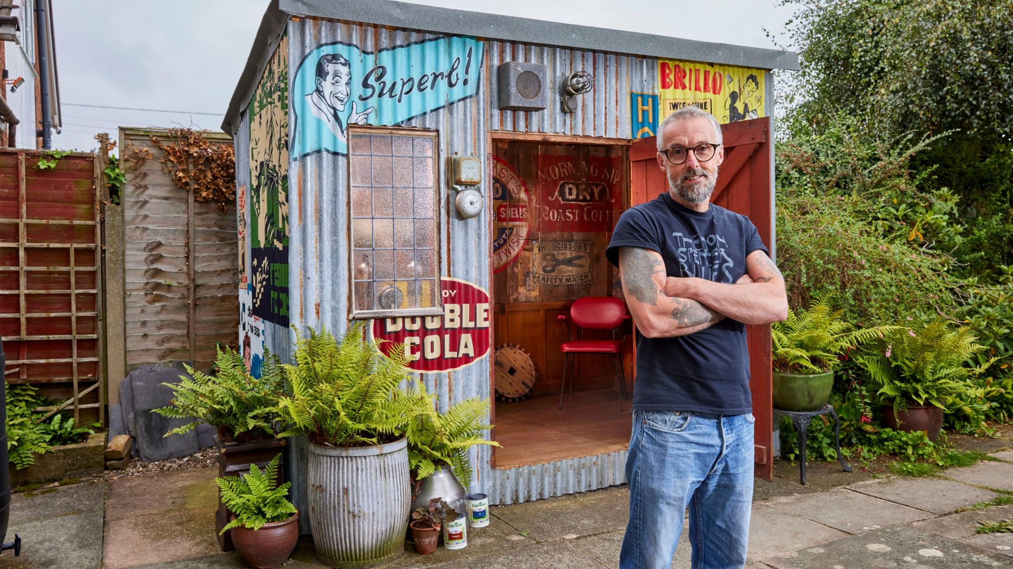 A man in a navy blue T-shirt and jeans stands in front of a corrugated shed with colourful painted slogans