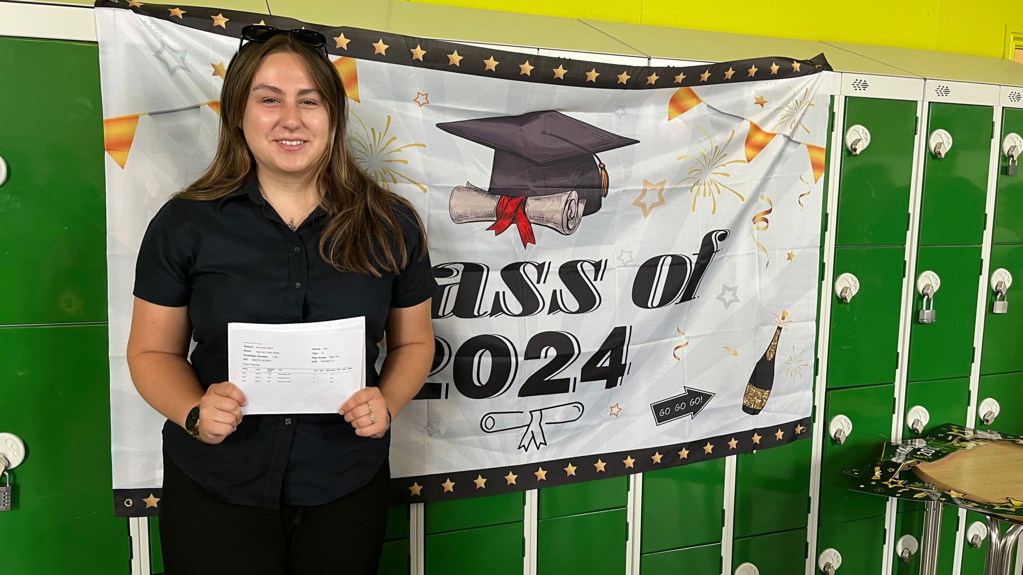 Hannah has long dark hair. She stands in front of a banner congratulating the class of 2024. She is wearing a plain black shirt and black skirt. She is holding her results paper in front of her. 