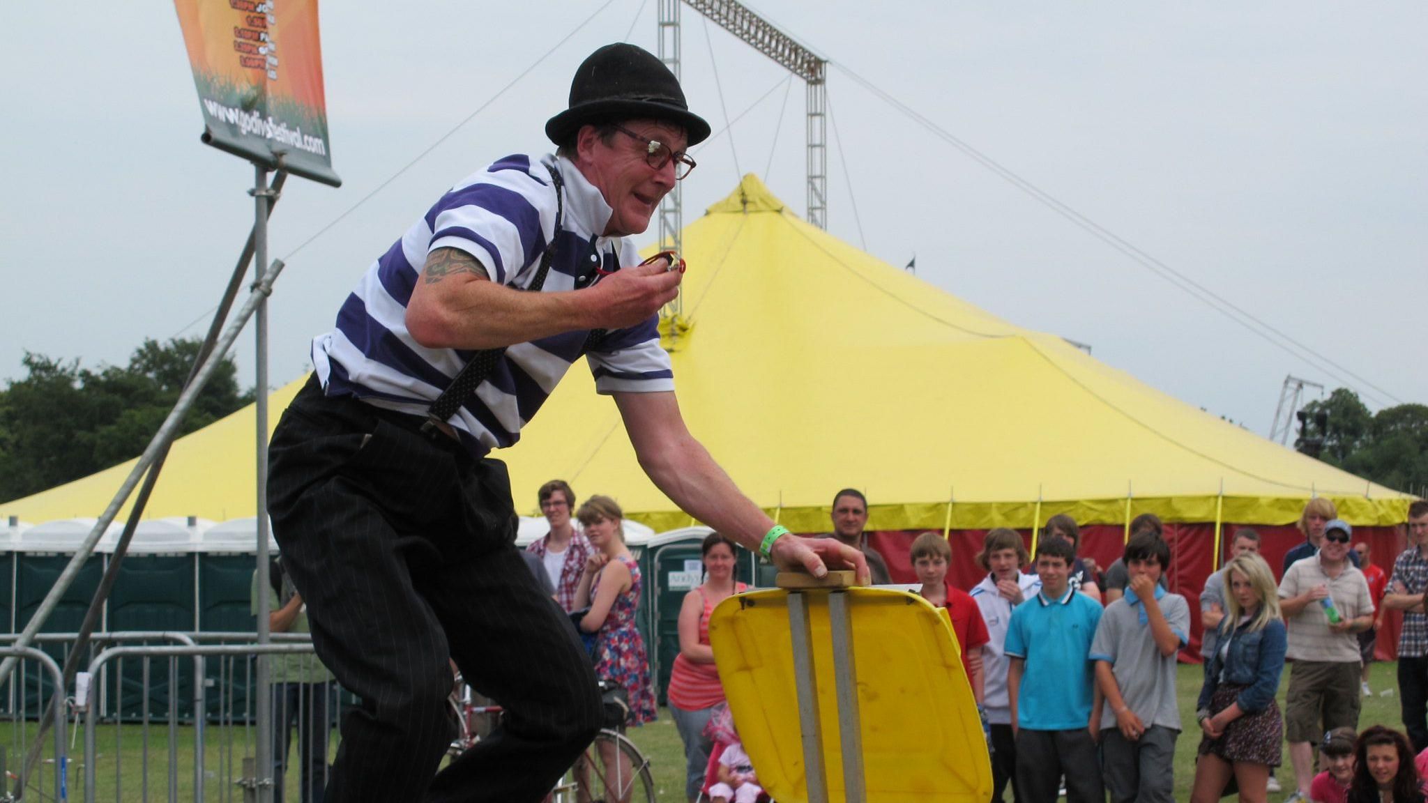 A clown at the festival in 2019. He is leaning forwards while his left hand is leaning on a yellow plastic square which appears to be anchored to the ground. He has a blue and white striped top with braces over the top connected to his black trousers, a black domed hat and glasses and is smiling. Behind him a small crowd can be seen including children and behind them is a large yellow tent