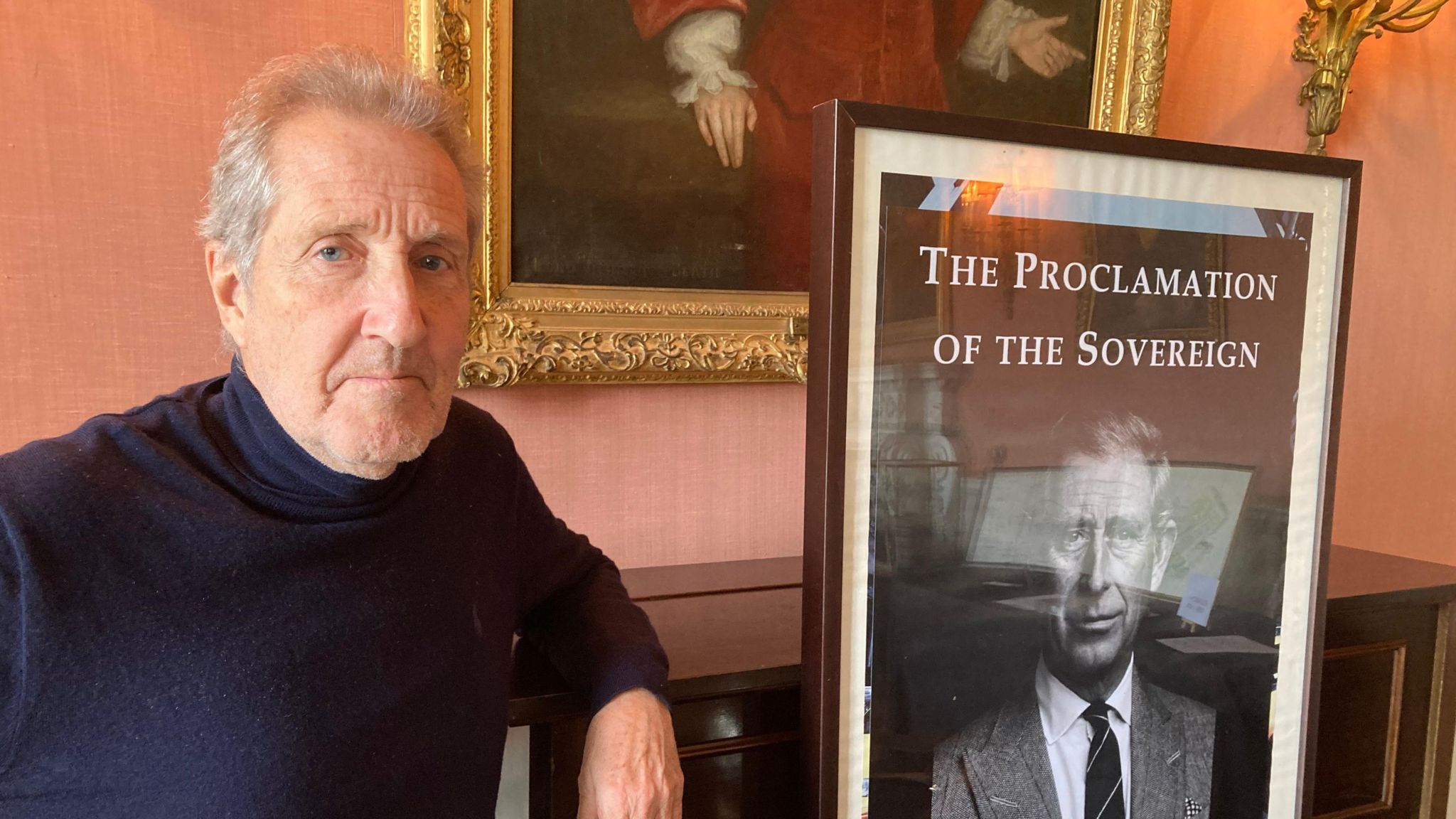 Man in a navy rollneck jumper stands next to a portrait of King Charles