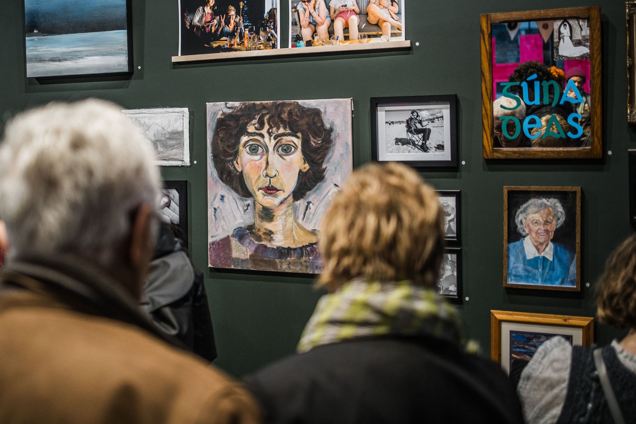 the backs of people's heads as they look at a crowded wall of art depicting people's mothers