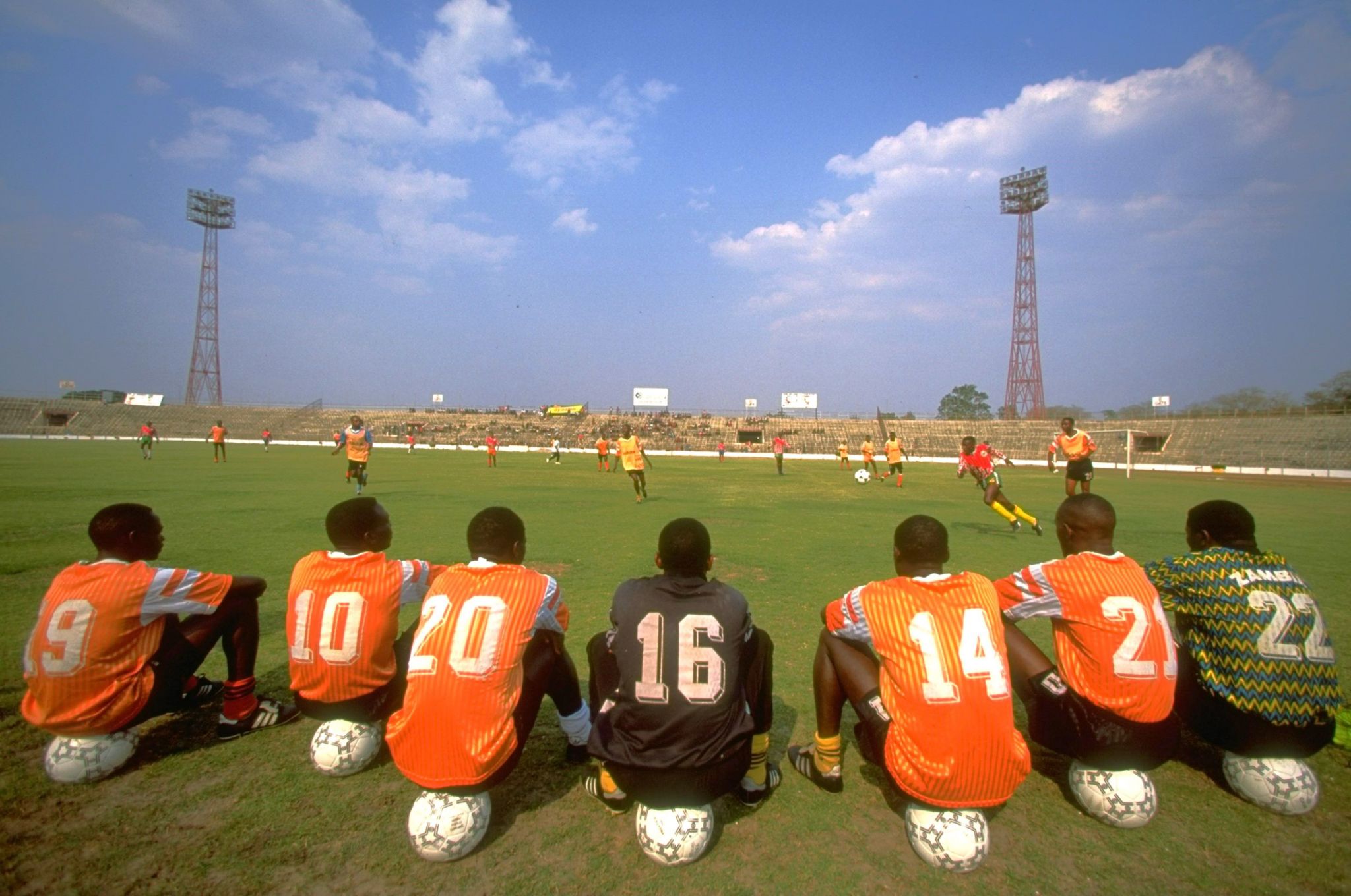 A group of orange-shirted footballers sit on footballs and watched a trial match taking place in the National Stadium in Lusaka