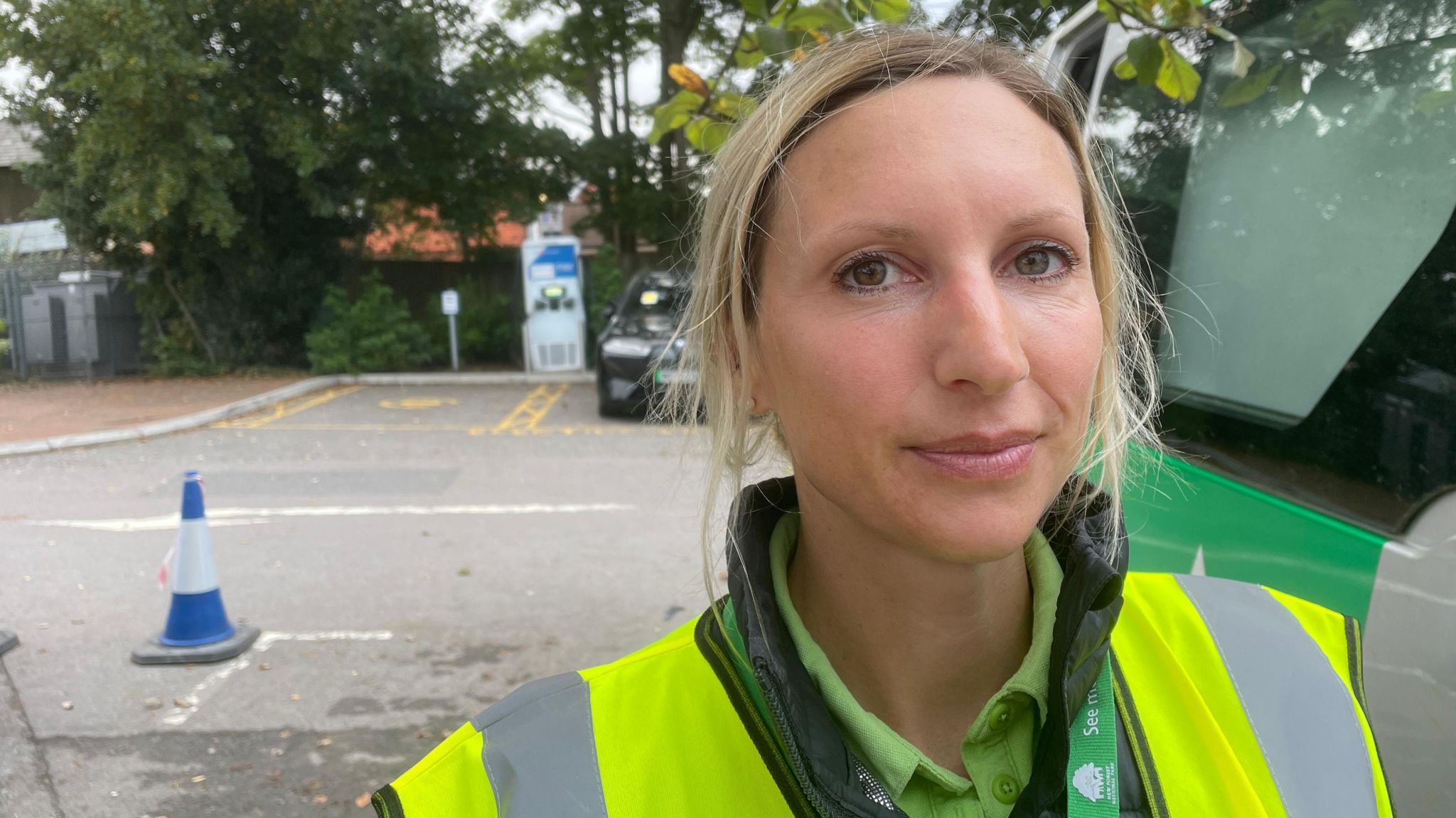 A woman is stood in a car park wearing a florescent jacket. She is stood next to a van and also wearing a green lanyard with the words 'Forestry England' on it. 