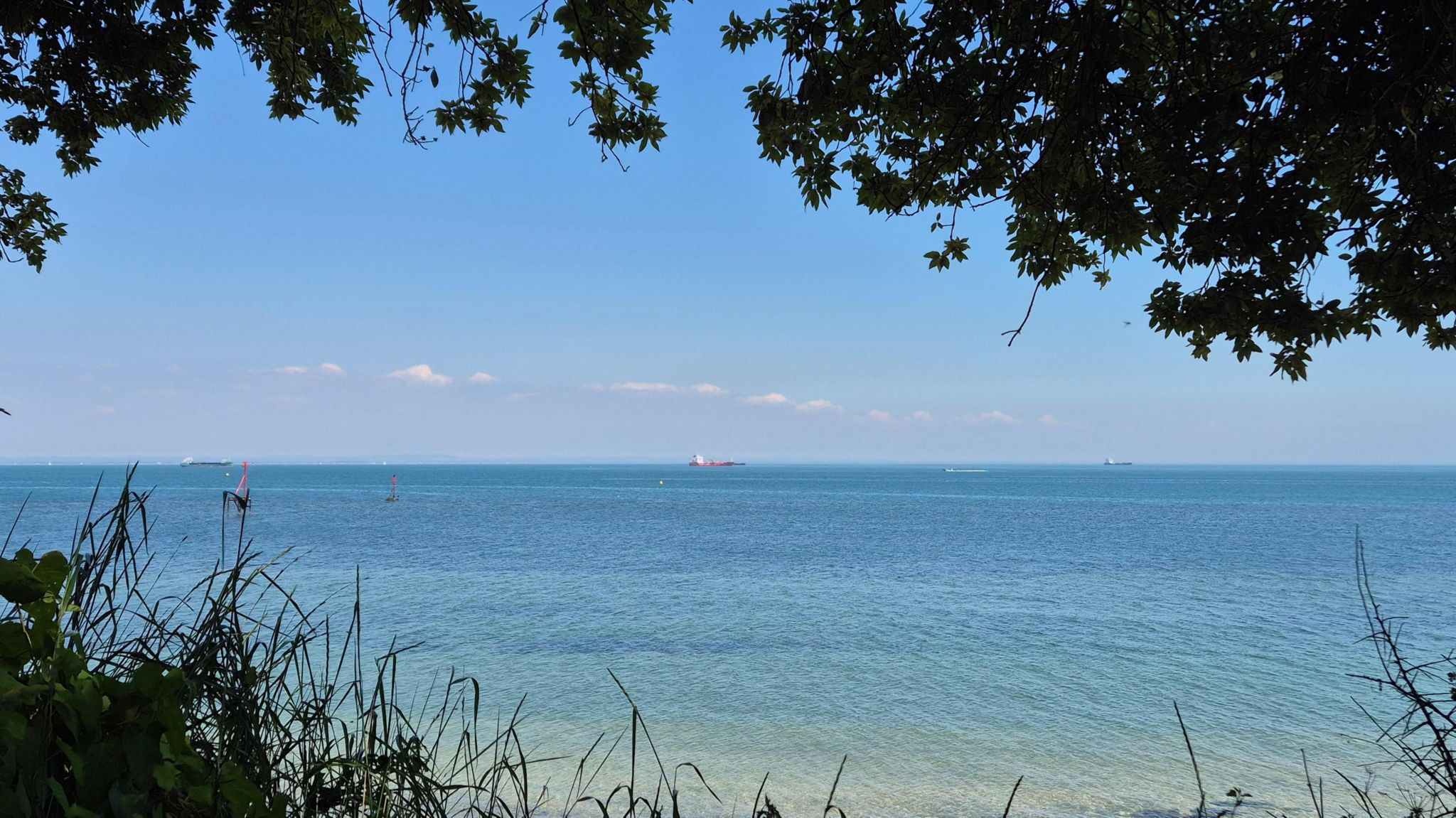A view of the bright blue sea on a sunny day with blue skies. Taken from Bembridge showing a boat on the horizon and with the picture framed by trees above and grass below