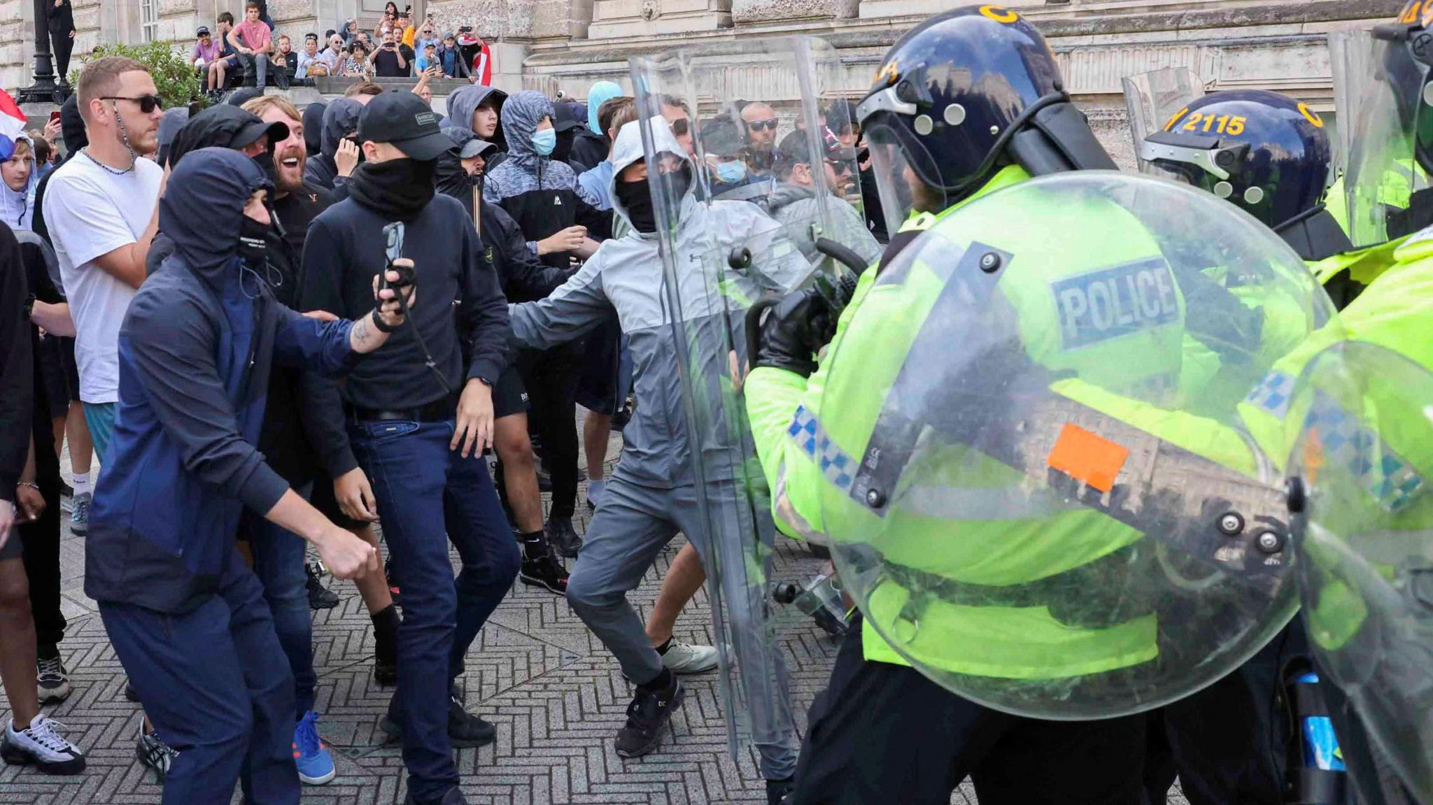 a group of police officers in riot gear and carrying clear shields push forward into a group of men in casual clothing, most of whom have their faces covered