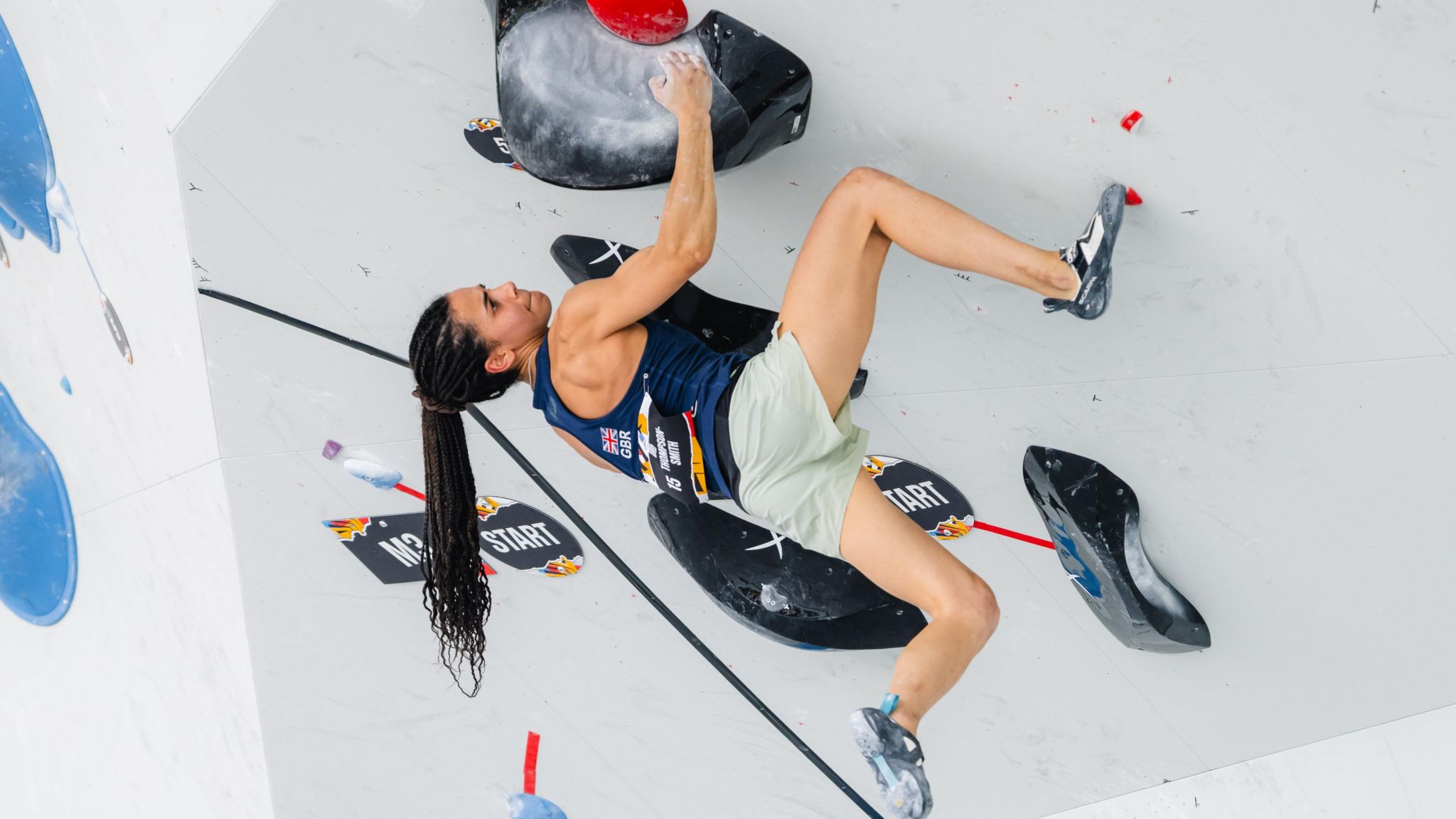 Molly Thompson-Smith climbing. Molly, in a Team GB vest and green shorts, clings to a climbing wall, one leg hanging loose. She has a determined look and her long ponytail hangs down. 