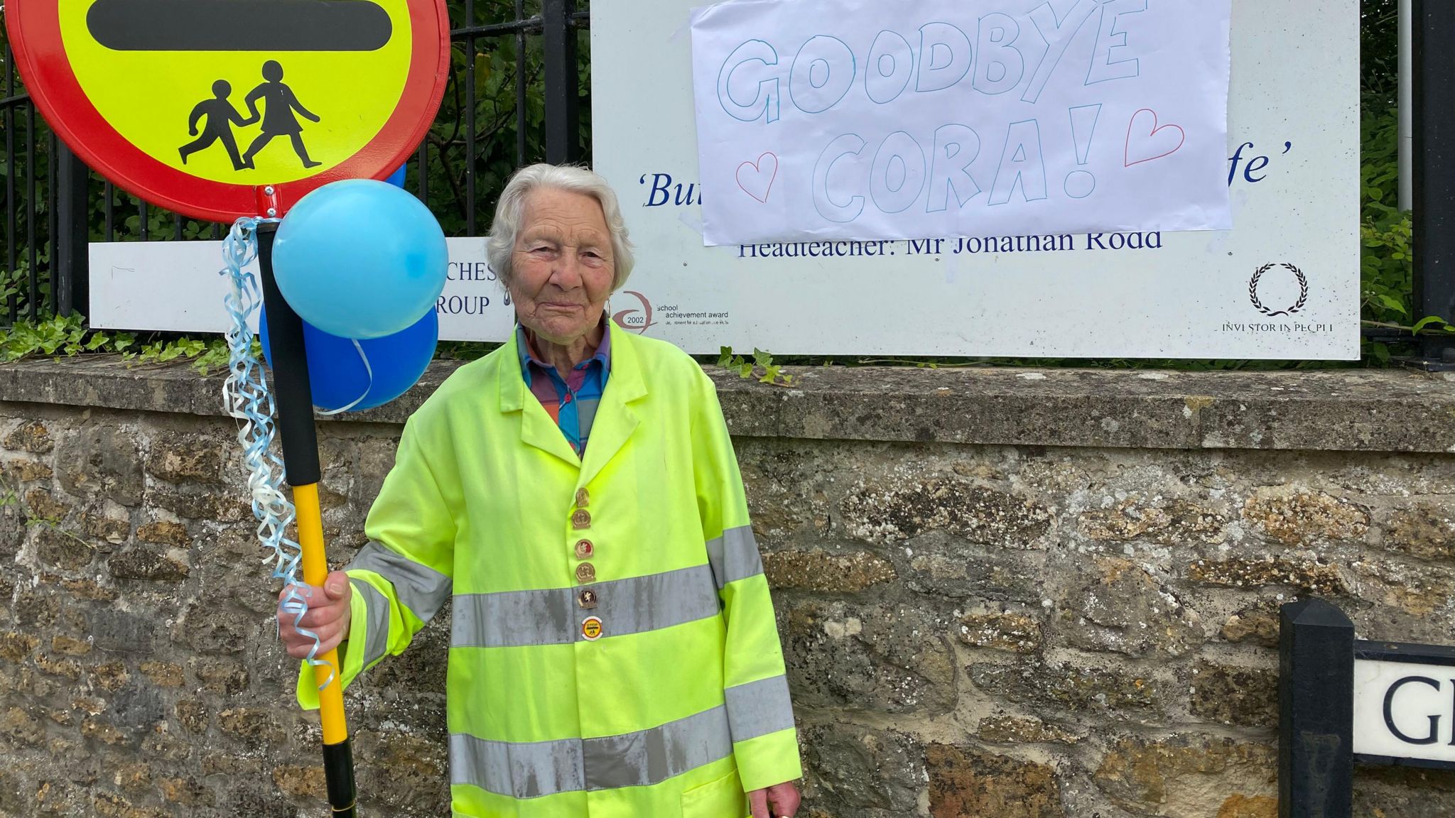 Cora standing in a hi-vis jacket as a lollipop lady holding a 'stop' sign with blue balloons on it