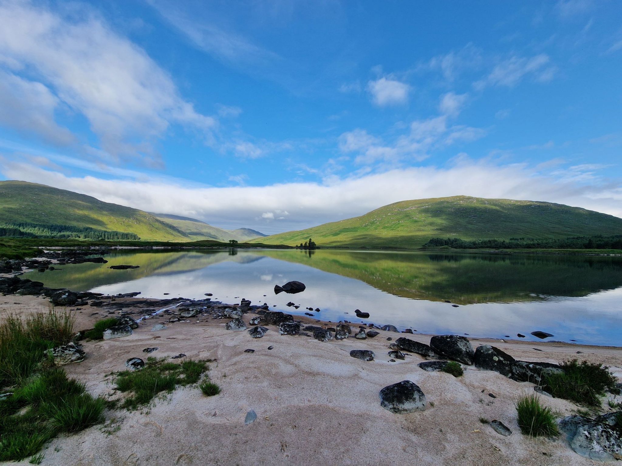 Landscape taken from a sandy beach looking out over clear water with two green hills in the distance and a very blue sky with few white clouds