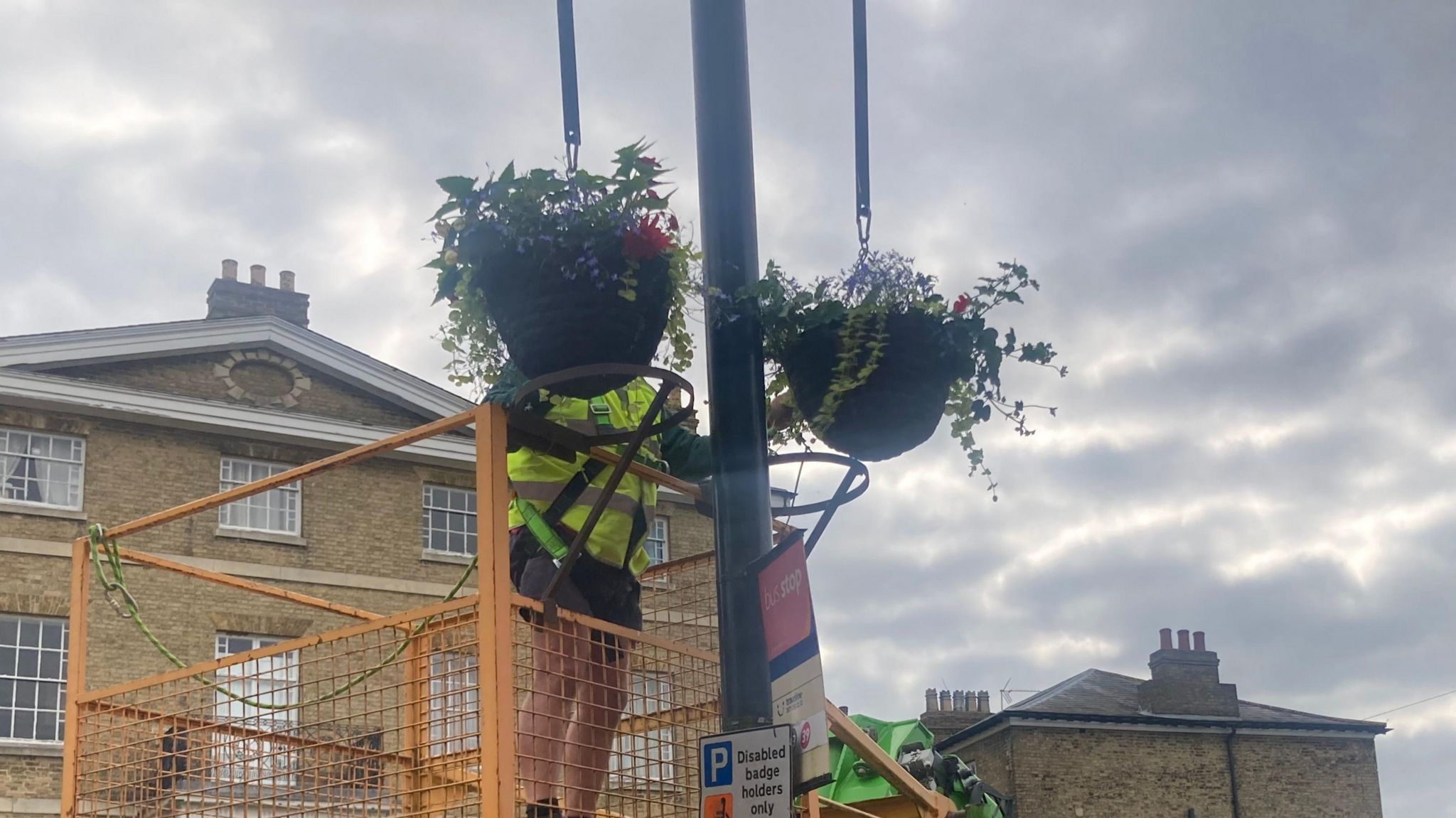 Hanging baskets being placed on street lamps by a person in a high-visibility jacket