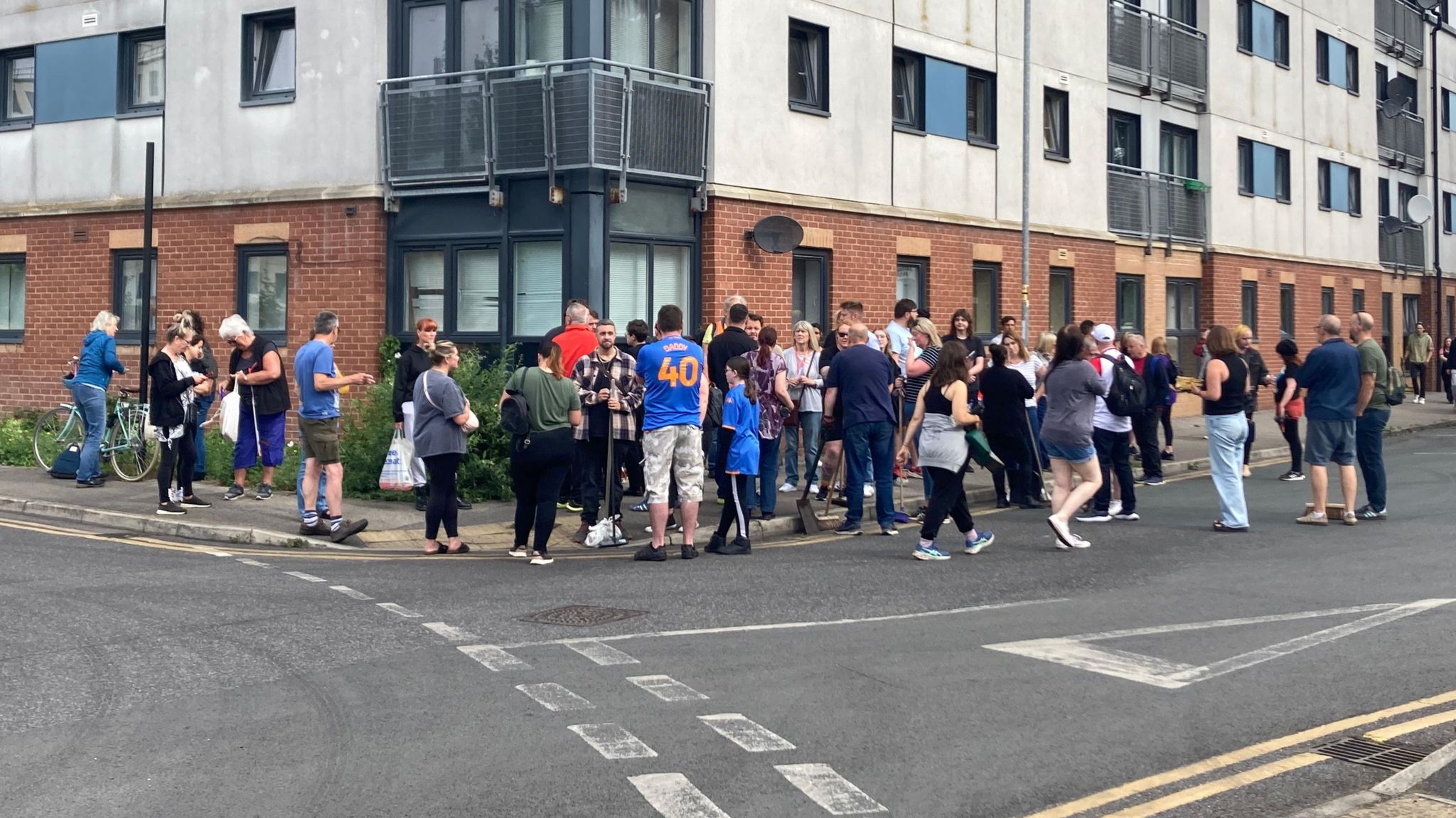 Members of the community stand on a street corner, some with brooms, ready to help with a clear-up operation. 