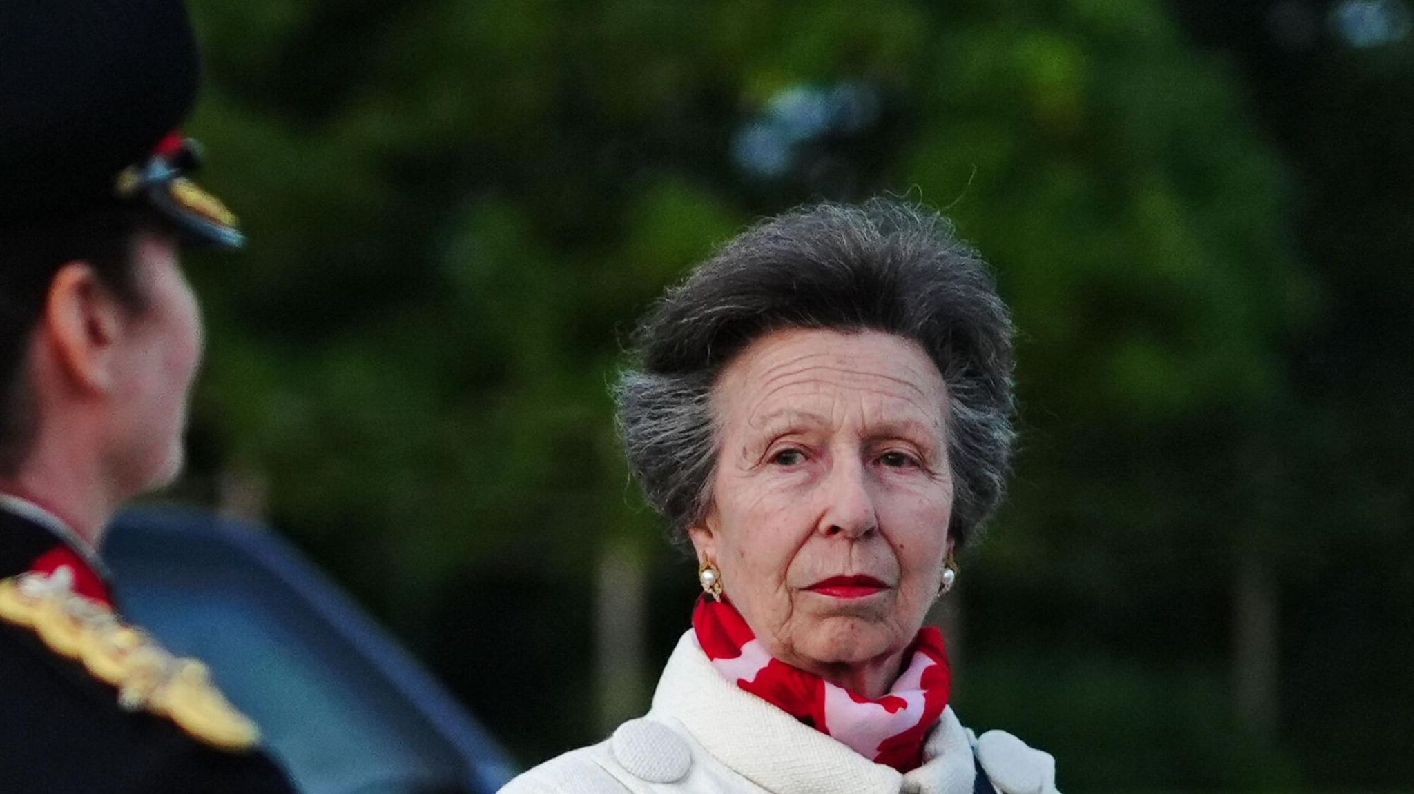 Princess Anne with red lipstick wearing white coat and pearl earrings.